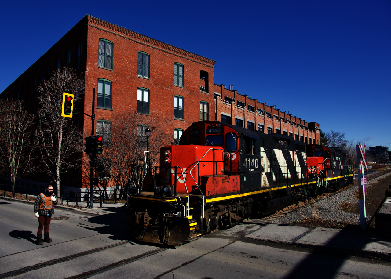 A pair of GP9's are returning to the main line after dropping off grain cars at Ardent Mills. They are crossing Charlevoix Street and a crewmember is flagging.