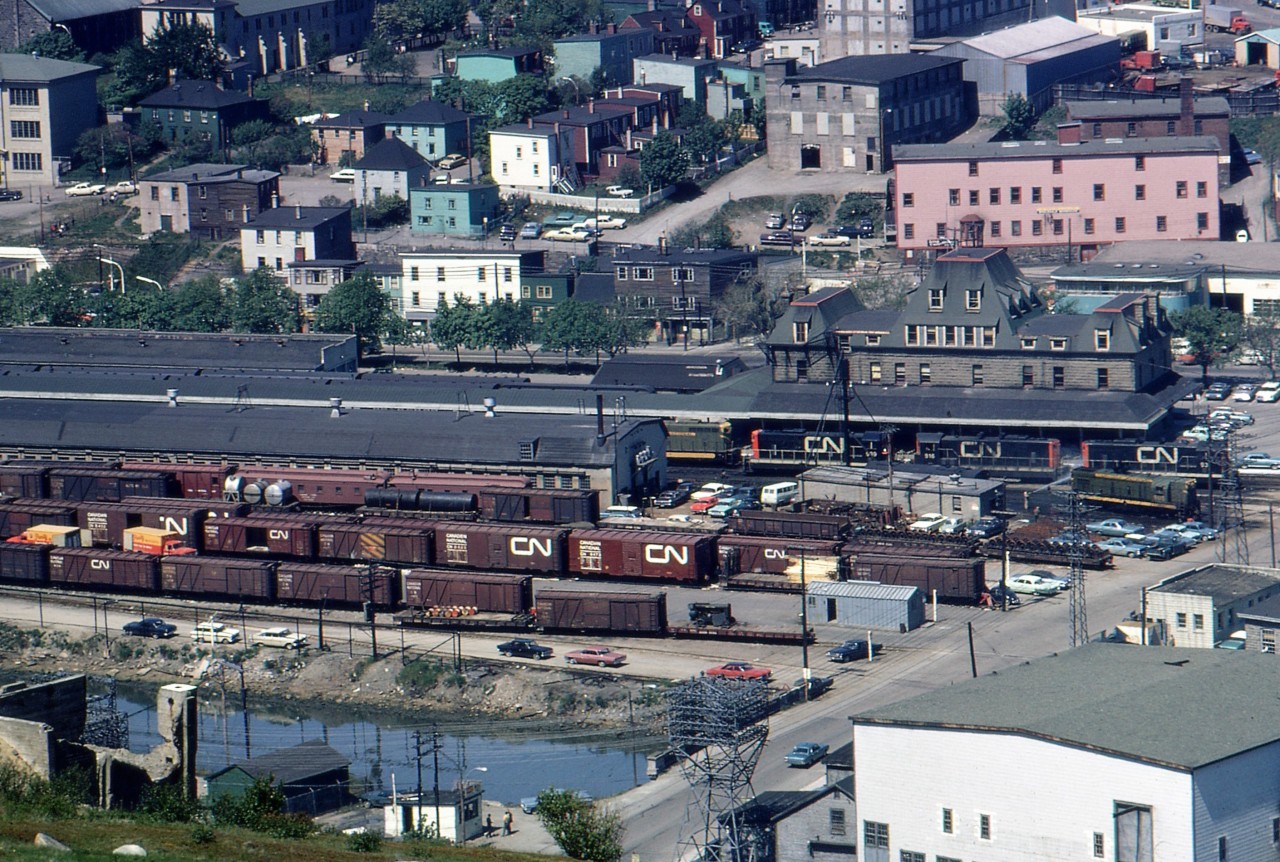 ST. JOHN'S SCENE - Travelling with four University of Toronto friends - including the late James A. Brown - to celebrate Canada's Centennial by riding Newfoundland's endangered passenger train 'Caribou', John Freyseng of Ontario was able to capture some amazing narrow gauge images. From high atop the South Side Hills, he photographs a downward view of the busy St. John's yard and the colorful homes after disembarking from a 22-hour overnight journey on CN Train No. 102, the eastbound 'Caribou'. The NF210 diesels are mostly repainted in the new noodle image but yet a handful will still remain in the old green and gold paint for another two years. The much smaller G8 to the right will have at least two of her five sisters wear the olive green until late 1978. The yard contains a mix of standard steel 'Mainland' 40-foot boxcars and the smaller narrow gauge outside braced ones, built specifically for Newfoundland operations. The Victorian era station, built with granite from the famous Gaff Topsails, opened in 1903 in an area that was reclaimed from the headwaters of the Waterford River and the St. John's Harbor. Once again, his friend Jim who while standing alongside him, captured pretty much an identical image. Today, nearly all is gone and the Pitts Memorial Highway Overpass intersects this scene but the station survives as the Railway Coastal Museum. The last scheduled train to pull into this yard was Terra Transport Extra 944 East on September 29, 1988. More of 1967 John Freyseng photos can be seen in my upcoming TRAINS OF NEWFOUNDLAND, to be released by Flanker Press on September 9, 2022.