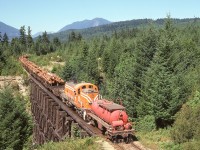 Less than a year after gaining its orange-with-white-stripes livery, Crown Zellerbach RS-3 4097 is eastbound with log loads from Nanaimo Lakes on the Boulder Creek bridge at 12.2 miles west of Ladysmith on Friday 1974-07-26.  Since it is fire season, the water car is continuously sprinkling the roadbed while a crew member has opened the soaker valve on the coupled end to minimize the chances of a bridge fire from brakeshoe sparks.