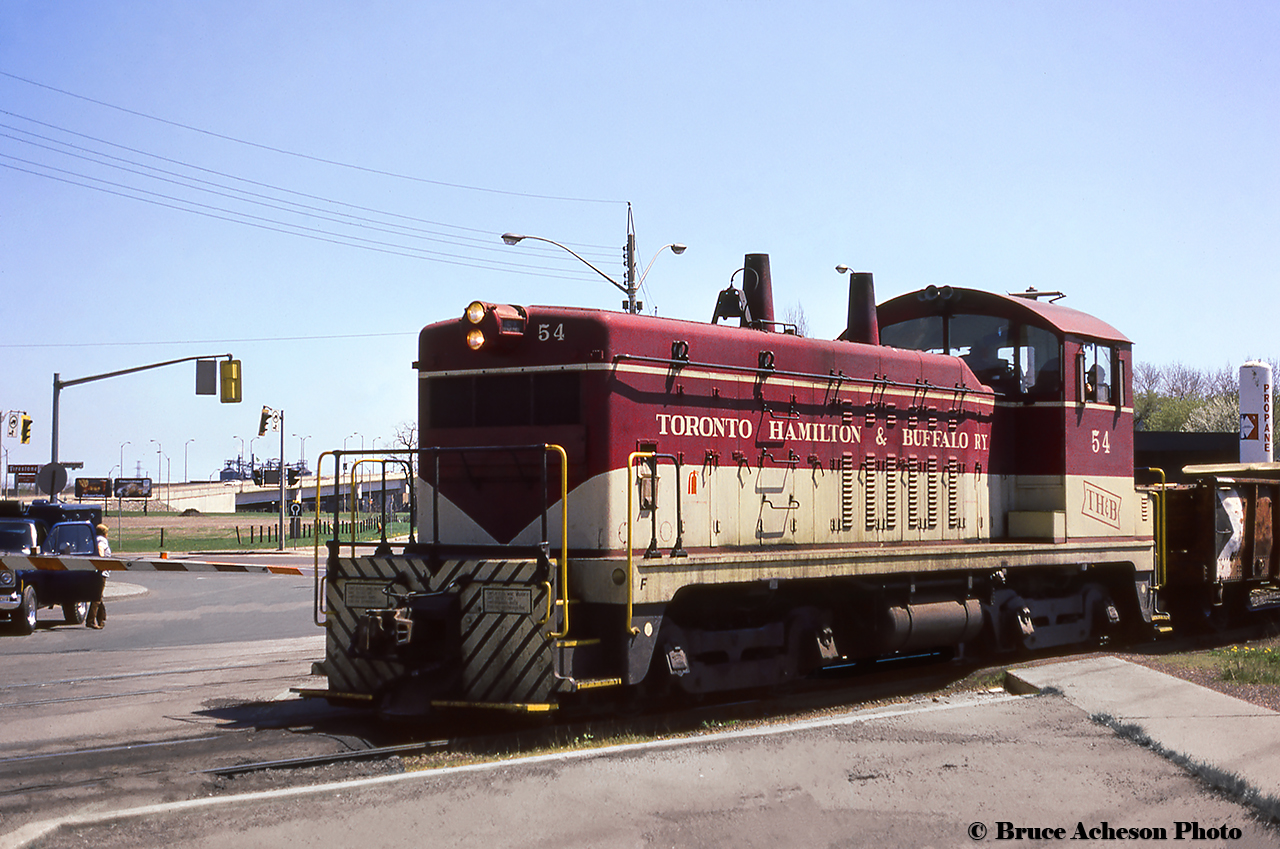 TH&B 54 leads a cut of cars across Kenilworth Avenue and Beach Road into Adams Yard.  In the background the on ramps for Nikola Tesla Boulevard can be seen climbing over Burlington Street and the Firestone Lead.  Note the Firestone signage near the plant at far left.