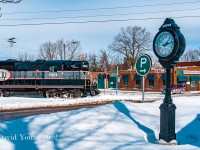 Having made a stop prior to occupying the crossing (note the stop sign), the circuit is triggered and the lights are activated for the crossing in downtown Stayner. The conductor can be seen keeping a watchful eye for any eastbound traffic along Highway 26 as the engineer throttles up and blows a "14L". This crossing was unique in the fact the signals were mounted on opposite sides of the road than usually seen (in this case mounted on the northside of the westbound lane but protecting the eastbound traffic and vice versa on the other side)- perhaps the reasoning was something to do with the side streets coming off their respective sides of the crossing? <br>

  In about thirteen hundred feet the conductor will be down, trudging through the snow as he works to <a href="http://www.railpictures.ca/?attachment_id=6942"> exchange the one loaded hopper in tow for the three empties on spot in the siding for F.S. Partners</a>.