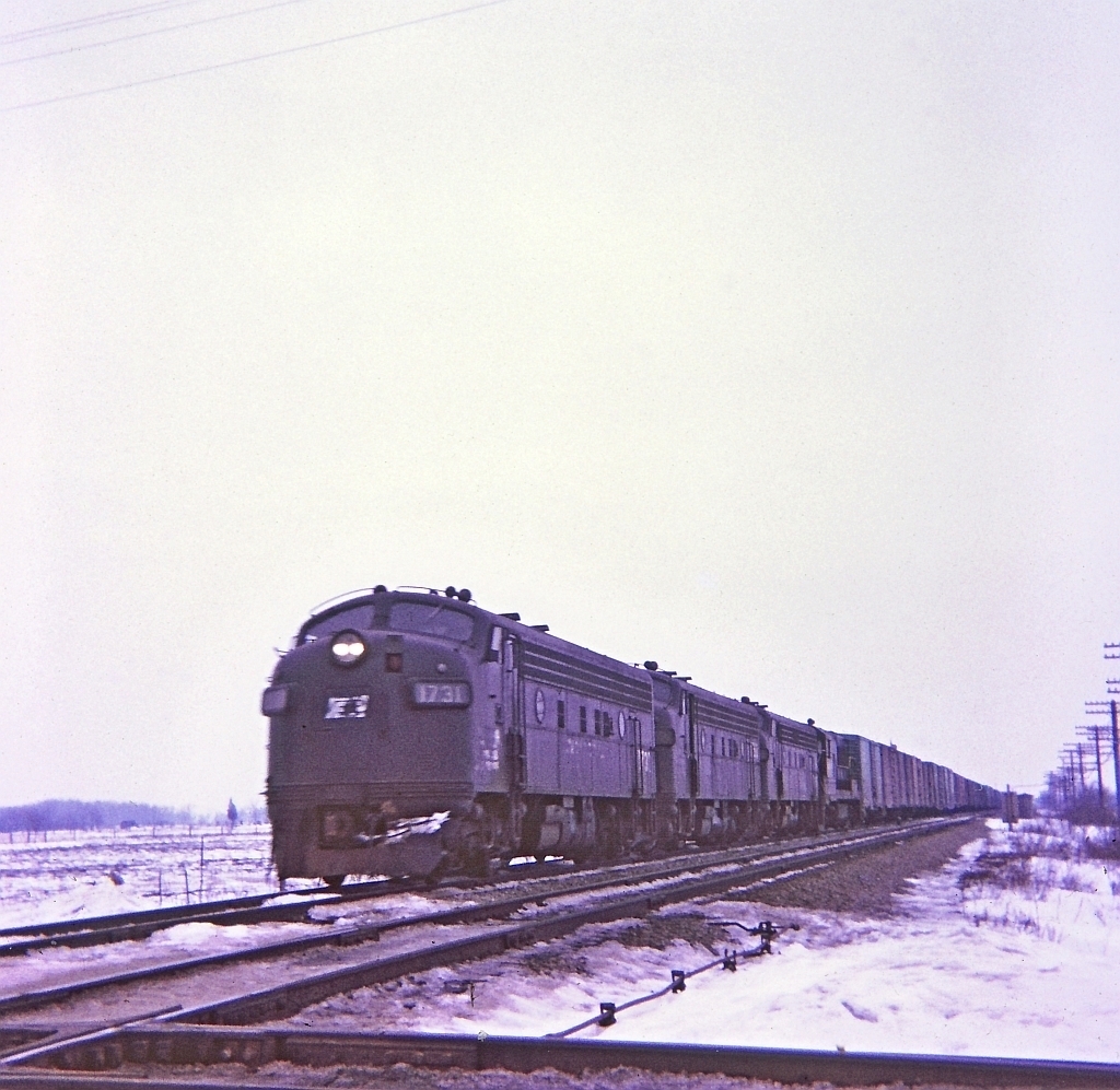 A dismal winter afternoon, even though this a color image might as well been black and white film. Train symbol MC-4 with a trio of F units and a U boat all elephant style running east on the double track CASO is about to bang over the diamond of the single track TH&B Dunnville sub. E&O tower was located at mile 30.5 on the Penn Central Canada Division CASO. A "Kodak Instamatic Moment" from long ago.