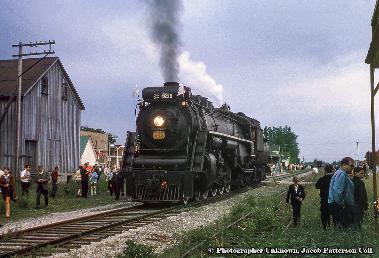 CNR 6218 pauses while headed eastbound during an excursion at Thedford on the Forest Subdivision. while crowds from the train, spotted at the station, surround the engine.  Part of the station can be seen beyond the tender.  Originally constructed as part of the Toronto – Chicago mainline of the Grand Trunk Railway in late 1856, the Forest Sub ran west from St. Mary’s Junction to Point Edward, just a couple miles north of the Great Western’s Sarnia yards.  The demise of the line came in sections, with Forest to Sarnia abandoned in 1982, Parkhill to Forest in 1986, and finally St. Mary[s junction to Parkhill in 1989.More shots along the Forest Sub:Calmachie, 1959, by Bill ThomsonForest, 1963, by Bill ThomsonOriginal Photographer Unknown, Jacob Patterson Collection Slide.