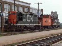 A gloomy summer day finds both of CN’s units assigned to Guelph parked in track XW41 across from <a href= http://www.railpictures.ca/?attachment_id=47517>the station.</a>  Finding both units at the station seems to have been an unusual occurrence as typically the larger unit, usually a GP9 by this time, would <a href= http://www.railpictures.ca/?attachment_id=49012>be based out of CN Guelph Junction</a> for use on the job to industries in Galt, while the SW1200RS or other similar power would be assigned to the yard job in town.  Power includes CN 4571, rebuilt to CN 7077 in November 1992 and still on the roster as of 2021.  CN 1229, retired 1992, to AMT HT-001, renumbered AMT 002, and finally Sidbec Feruni as their 002.  Disposition unknown.<br><br>Beyond Wyndham Street, the battlements of the Guelph Armoury, built in 1906, can be seen, still in use today.  Built in the early 1900s and for years part of the scene greeting travelers arriving at the station, the factory at left was in use by Guelph Cotton Mills per a 1907 fire insurance map, and by 1908, taken over by the Louden Machinery Company for the manufacturing of agricultural equipment. The Aspinwall Manufacturing Company would assume operation of the site in 1912 as their only Canadian plant <a href= https://s3.amazonaws.com/pastperfectonline/images/museum_51/091/2009321996.jpg >(seen here circa 1915)</a> making equipment for potato farmers, including automated potato planters, sprayers, cutters, sorters, and weeders.  Per a 1929 fire insurance map, the property was part of the textile industry under the Regent Knitting Company, and later by Zephyr Looms and Textiles Inc. in the late 1930s. Zephyr Looms & Textiles worked through the Second World War handling military contracts for uniform articles and other small items. Emerging from the war, the company would rebrand as Textile Industries Ltd. in December 1945, which it would operate as until closing for good in December of 1980. At some point during the 1970s a slight change in the name recognized their location in Guelph on Wyndham Street, as they became known as <a href= https://s3.amazonaws.com/pastperfectonline/images/museum_51/061/199228190.jpg>Wyndham Textile Industries Limited.  The building was demolished in the early 2010s to make way for GO Transit parking.</a>.<br><br><i>Original Photographer Unknown, Jacob Patterson Collection Slide.</i>
