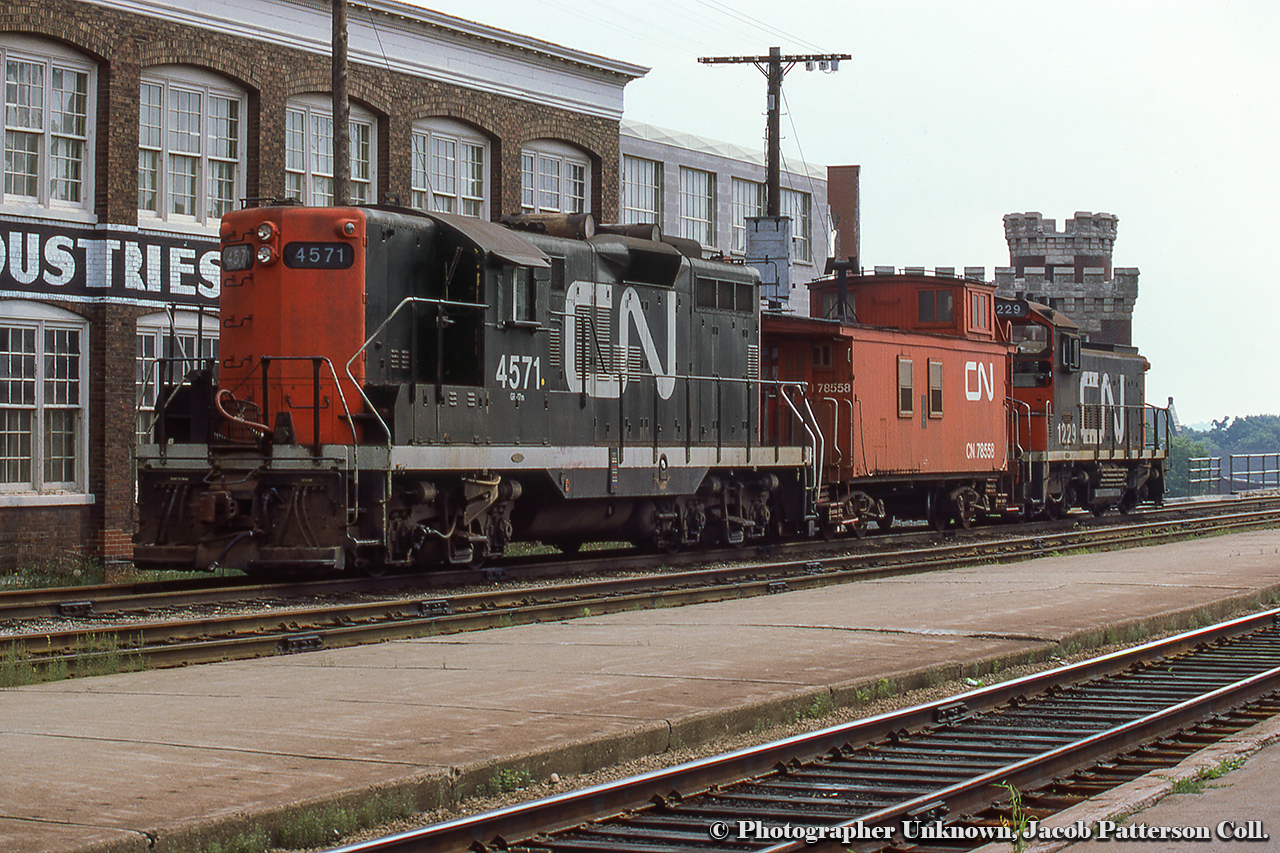 A gloomy summer day finds both of CN’s units assigned to Guelph parked in track XW41 across from the station.  Finding both units at the station seems to have been an unusual occurrence as typically the larger unit, usually a GP9 by this time, would be based out of CN Guelph Junction for use on the job to industries in Galt, while the SW1200RS or other similar power would be assigned to the yard job in town.  Power includes CN 4571, rebuilt to CN 7077 in November 1992 and still on the roster as of 2021.  CN 1229, retired 1992, to AMT HT-001, renumbered AMT 002, and finally Sidbec Feruni as their 002.  Disposition unknown.Beyond Wyndham Street, the battlements of the Guelph Armoury, built in 1906, can be seen, still in use today.  Built in the early 1900s and for years part of the scene greeting travelers arriving at the station, the factory at left was in use by Guelph Cotton Mills per a 1907 fire insurance map, and by 1908, taken over by the Louden Machinery Company for the manufacturing of agricultural equipment. The Aspinwall Manufacturing Company would assume operation of the site in 1912 as their only Canadian plant (seen here circa 1915) making equipment for potato farmers, including automated potato planters, sprayers, cutters, sorters, and weeders.  Per a 1929 fire insurance map, the property was part of the textile industry under the Regent Knitting Company, and later by Zephyr Looms and Textiles Inc. in the late 1930s. Zephyr Looms & Textiles worked through the Second World War handling military contracts for uniform articles and other small items. Emerging from the war, the company would rebrand as Textile Industries Ltd. in December 1945, which it would operate as until closing for good in December of 1980. At some point during the 1970s a slight change in the name recognized their location in Guelph on Wyndham Street, as they became known as Wyndham Textile Industries Limited.  The building was demolished in the early 2010s to make way for GO Transit parking..Original Photographer Unknown, Jacob Patterson Collection Slide.