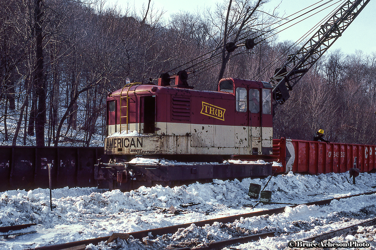 TH&B's self-propelled American Hoist and Derrick X-755 is seen equipped with a clamshell bucket at Kinnear Yard while working to clear snow from the track for maintenance.  It appears some minor derailment cleanup is underway with wood cribbing scattered around and rails rolled to the side.