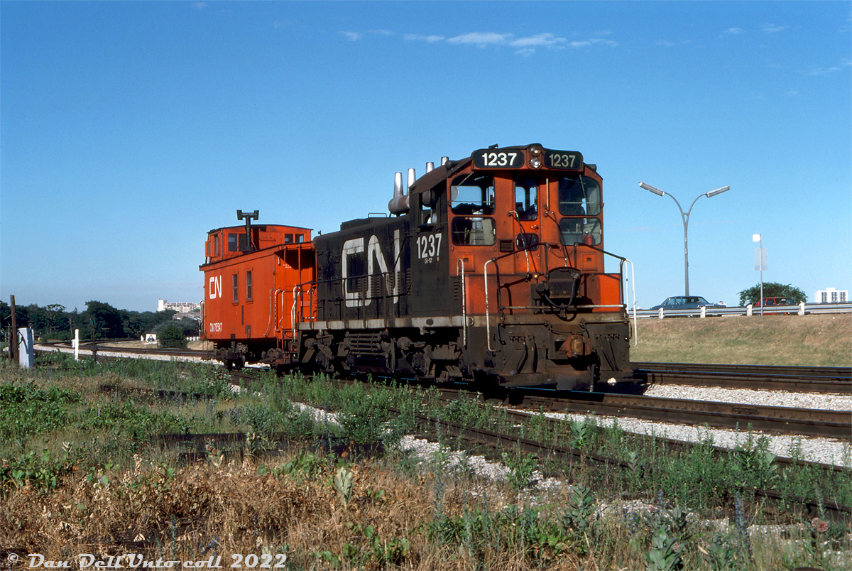 Running parallel to the Gardiner Expressway, CN SW1200RS 1237 trundles westbound at Mile 4.7 on CN's Oakville Sub on a "caboose hop" with wooden van 78547 on a local job (possibly heading to switch the nearby Ontario Food Terminal, or returning light power to Mimico Yard.

In the foreground is the switch to the former CN Humber Belt Line Spur, once the western branch of the Toronto Beltline Railway, but by this time a siding to the nearby Stelco Swansea Works at The Queensway & Windermere. The Toronto Beltline's "Humber Loop" once ran from here northward and eastward to West Toronto in the 1890's, but was cut down to small spurs when CN predecessor Grand Trunk Railway took over and began running freight. In the 1950's The Queensway was extended west and the spur (still serving one or two customers to the north) had diamonds and crossing protection installed for streetcars. This setup lasted until removal in the mid-60's. Around that time, Stelco expanded their property south over part of the line, and a small portion branching from the Oakville Sub here served as a siding for Stelco into the 1990's (the factory opened in 1885, closed in 1990, demolished for redevelopment in the early 2000's).

Charles Houser photo (maybe), Dan Dell'Unto collection slide.