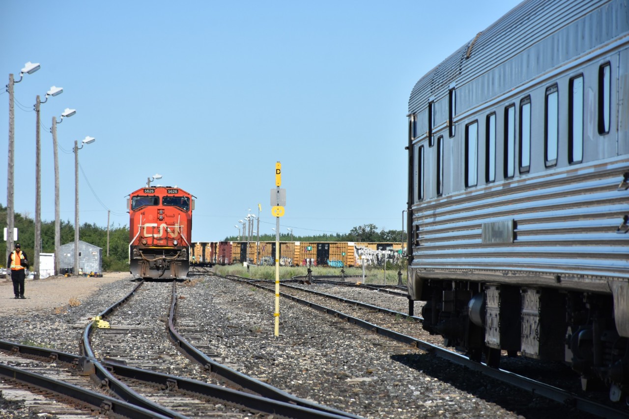 Assiniboine Jct. Mile 0.00 on CN's Assiniboine Sub. is a busy place this morning with CN 5626 sitting idle in the yard waiting for its next assignment, VIA 692 (VIA 8229 Chateau Viger sleeping car) holding the main in front of the Canora, SK station, and a moving CN freight in the background. Other interesting sights in the photo are a conductor walking back to the station, tall yard lighting poles, derails, restricted clearance sign, wind sock, station name sign, mile board, and some switch stands, several with unique targets.