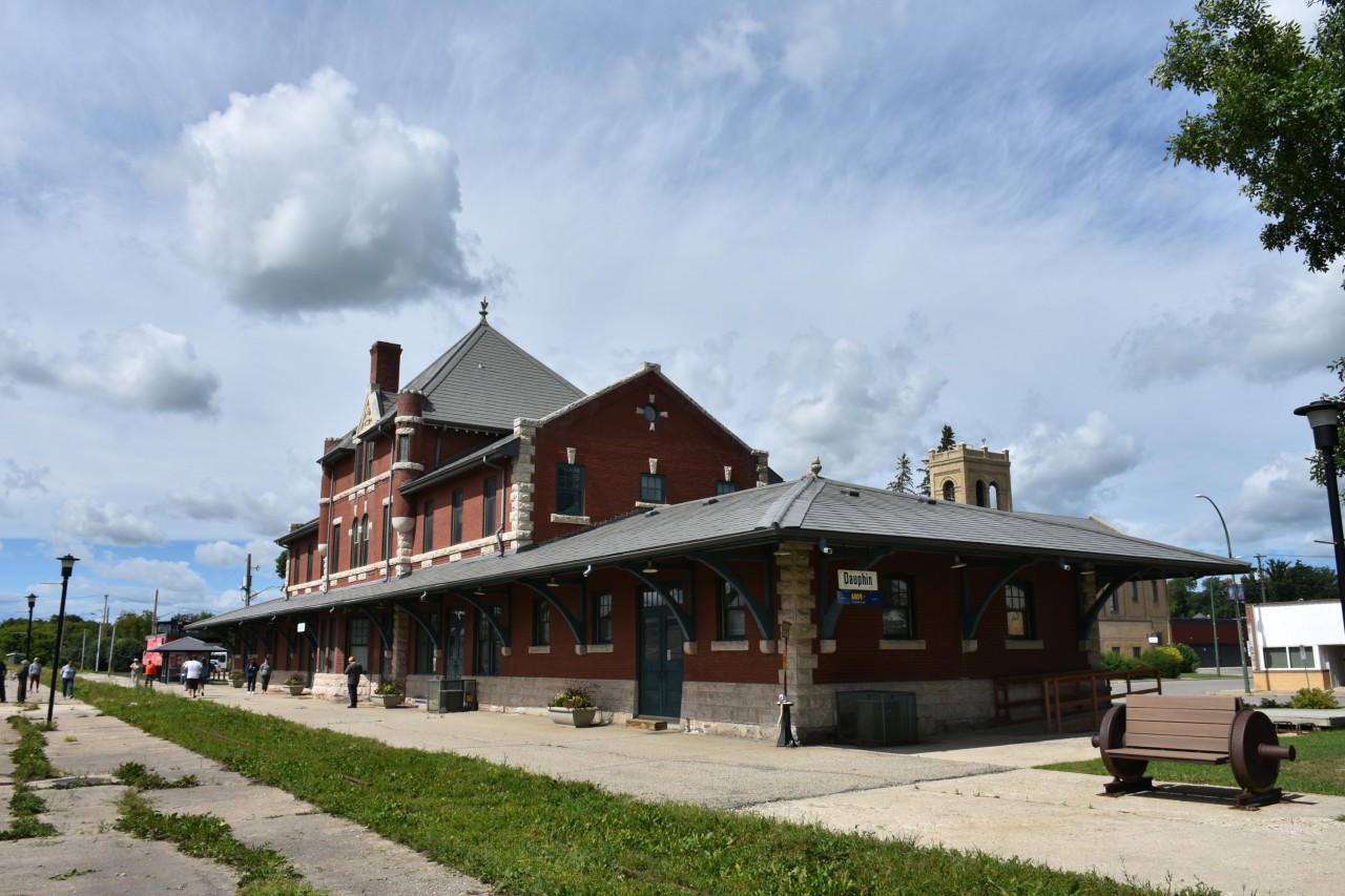 This mid-afternoon shot of VIA's Dauphin, MB station has a few passengers out on the platform stretching their legs during another stop on the 48 1/2 hour journey from Churchill to Winnipeg. Platform benches made from old railcar wheelsets dot this end of the station grounds, while a retired CN caboose (CN 79727) and a piece of maintenance equipment are on display at the opposite end. Dauphin station is home to the Dauphin Rail Museum. A view from the opposite end of the platform can be seen here;
http://www.railpictures.ca/?attachment_id=49621