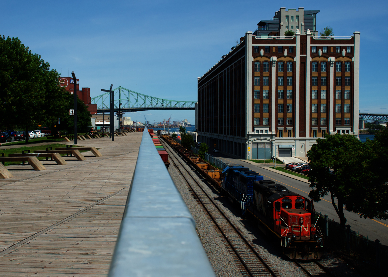 The Pointe St-Charles Switcher is passing an observation deck that gives a good view of the western end of the Port of Montreal as it doubles its train together. At right is a cold storage warehouse that has been converted to condos.