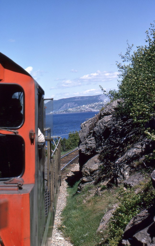 CARIBOU CLINGING TO CLIFFS - Continuing eastward with four University of Toronto friends - including the late James A. Brown - to celebrate Canada's Centennial by riding Newfoundland's endangered passenger train 'Caribou', John Freyseng of Ontario was able to capture some amazing narrow gauge images. Looking forward from his unique vantage point in second unit NF210 911, he photographs CN Train No. 102, the eastbound 'Caribou' rounding the high cliffs above the Humber Arm of the Bay of Islands just east of Curling. With the Humbermouth area of the City of Corner Brook in the background, Engineer Kevin Byrne and Fireman Austin Bennett up ahead will take the 919, 911 and the rest of the consist to its next stop in Corner Brook for a crew change in Newfoundland's second city. With the expert driving of Engineer Byrne, seen here navigating his train around rocks that are just inches away from the cab window, the train will descend to nearly sea level just ahead. More of John Freyseng's 1967 photos, as well as James A. Brown's, can be seen in my just released TRAINS OF NEWFOUNDLAND, from Flanker Press in both hard and softcover editions.