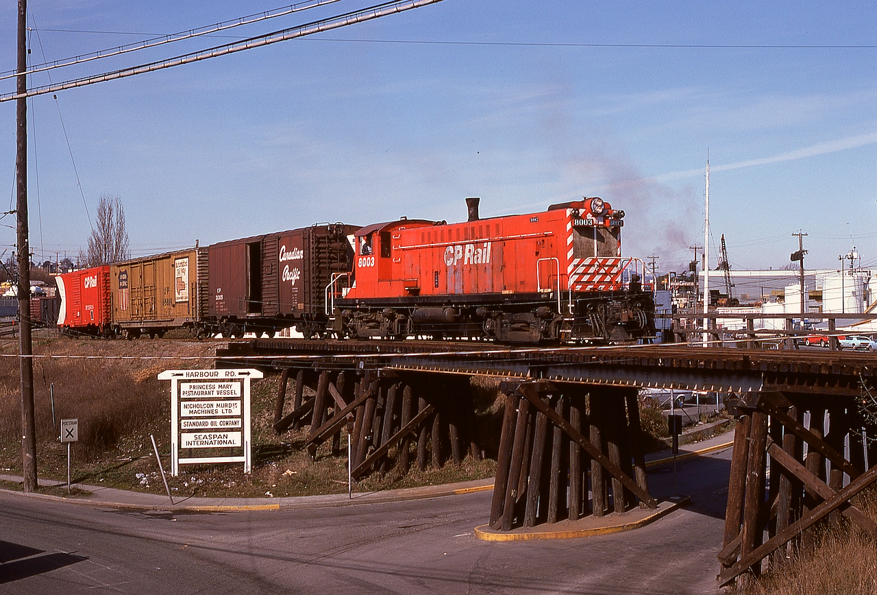 Occasionally, typically to verify some just-completed maintenance such as traction motor changeout was okay, the 0600 yard crew at Victoria was provided a Baldwin roadswitcher instead of the usual MLW S-3, and on Thursday 1975-03-06 that was the assignment for 8003.  Immediately north (by timetable, west by compass) of the Johnson Street bridge over Victoria harbour, a spur connected down a grade to an interchange with CN, and in this shot the yard crew is returning upgrade and crossing over the Harbour Road bridge just before reaching the main track switch.  This photo was taken from an overpass of Esquimalt Road that is today’s end of track at mileage 0.2.

Completely unknown to me at that time, it was only five days before shutdown of all seven remaining Baldwin roadswitcher units was begun, so this was a happy coincidence of sunshine and blind luck timing and my camera if ever there was one.