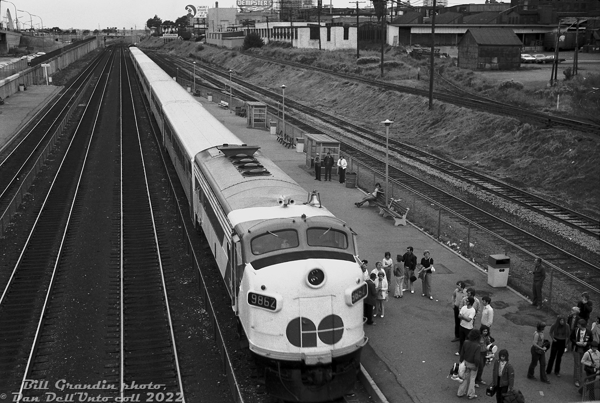 CNE season is in full swing as GO Transit APCU 9862 trails a westbound Lakeshore line commuter train out of Exhibiton GO Station. Passengers make their way to the overhead pedestrian bridge to cross the tracks south to the CNE (Canadian National Exhibition, held seasonally every year for a few weeks in August-September), as others stand around having a smoke.

The train in on CN's busy Oakville Sub west of downtown Toronto in the industrial Liberty Village district. Visible to the north along the station platforms is a small yard (some references call it CN's Strachan Avenue yard), many old factory buildings, and some of the sidings and industrial leads. A Penn Central boxcar is spotted on the siding for Central Warehouse. The long industrial lead on the incline connects to the Mowat Avenue Lead, and curves north along the side of Mowat to more industrial sidings around Liberty Street (including for Toronto Carpet Manufacturing, and GE's lamp factory). Dempster's (Canada Bread) had a bakery in the distance for many decades until closing in the early 2010's.

GO 9862 (originally converted from Ontario Northland FP7 1513 into a cab control/HEP unit for the startup of GO's Georgetown line in 1974) would be renumbered to 904 next year, retired and sold to Tri-Rail in 1991, and eventually end up at the Gold Coast Railroad Museum.

Bill Grandin photo, Dan Dell'Unto collection negative.

Alternate angles of Exhibiton station:
http://www.railpictures.ca/?attachment_id=46613
http://www.railpictures.ca/?attachment_id=35906