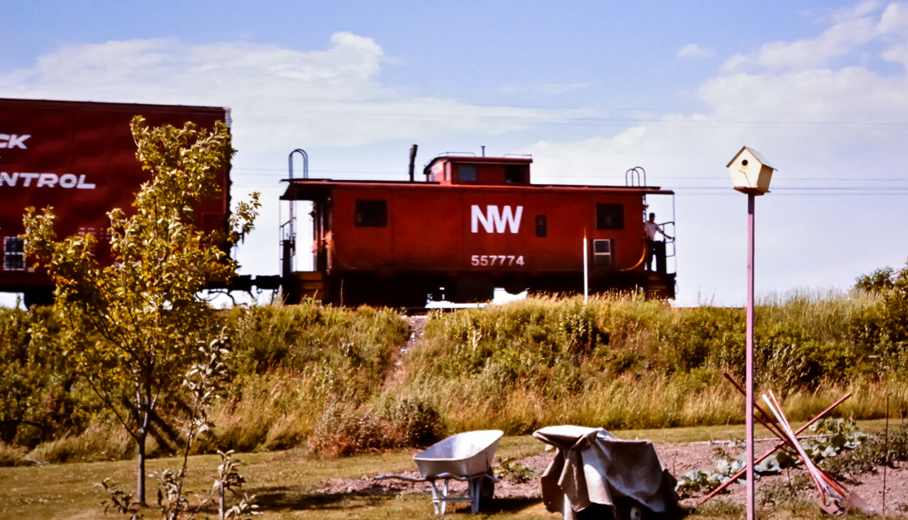 With the head end captured in a previous post, http://www.railpictures.ca/?attachment_id=50588 here is the tale end of the same westbound N&W freight scooting along the Cayuga Sub. through Jarvis ON in the summer of 1976.  Caboose NW 557774 is carrying the markers today. Just like the headend crew, one of the tail end crew members stands on the rear platform ready to acknowledge any of us that might be out in my parents back yard. The west backtrack switch, the restricted clearance sign for the elevated freight shed platform at the mill, & the pole line wires are clearly visible along the right-of-way. The wheelbarrow and garden tools by the well tilled garden belong to my dad, a.k.a. the CN Roadmaster stationed in Jarvis at the time. My parents house was less than a hundred feet from the Cayuga Sub. which, unknown to us at the time, was a treasure trove of railroad action waiting to be photographed. That worn path up the side of the hill now leads to a much larger trail where the Cayuga Sub. once was. Norfolk & Western is no more. The Cayuga Sub. is no more. And the garden is no more. :-( My how things change!