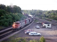 Back in the days when gatherings at the Junction were permitted, often with visits from the local police just to say "hello" and ask if there were any problems, summer nights were most enjoyable.
Not sure of the time captured here, but looking at the shadows thrown by the automobiles, it must be coming up to 2030 or so.  A nice catch was westbound CN freight with 3 GP38-2s for power; CN 5570, 5508 and 5515. (In 1988 these were renumbered to 4770, 4708 and 4715.)
Over by the "shack" watching the train pass is a young and up & coming talented photographer Dave Stowe with his long-time friend Bill McArthur, who was rather legendary around these parts over a 40 plus year span of shooting trains at Bayview. Bill has passed on; the junction ground is neglected and quiet. A fence sees to that. An era faded into history, but most fondly remembered.