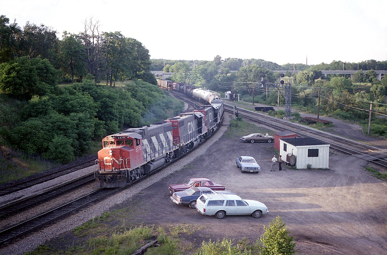 Back in the days when gatherings at the Junction were permitted, often with visits from the local police just to say "hello" and ask if there were any problems, summer nights were most enjoyable.
Not sure of the time captured here, but looking at the shadows thrown by the automobiles, it must be coming up to 2030 or so.  A nice catch was westbound CN freight with 3 GP38-2s for power; CN 5570, 5508 and 5515. (In 1988 these were renumbered to 4770, 4708 and 4715.)
Over by the "shack" watching the train pass is a young and up & coming talented photographer Dave Stowe with his long-time friend Bill McArthur, who was rather legendary around these parts over a 40 plus year span of shooting trains at Bayview. Bill has passed on; the junction ground is neglected and quiet. A fence sees to that. An era faded into history, but most fondly remembered.