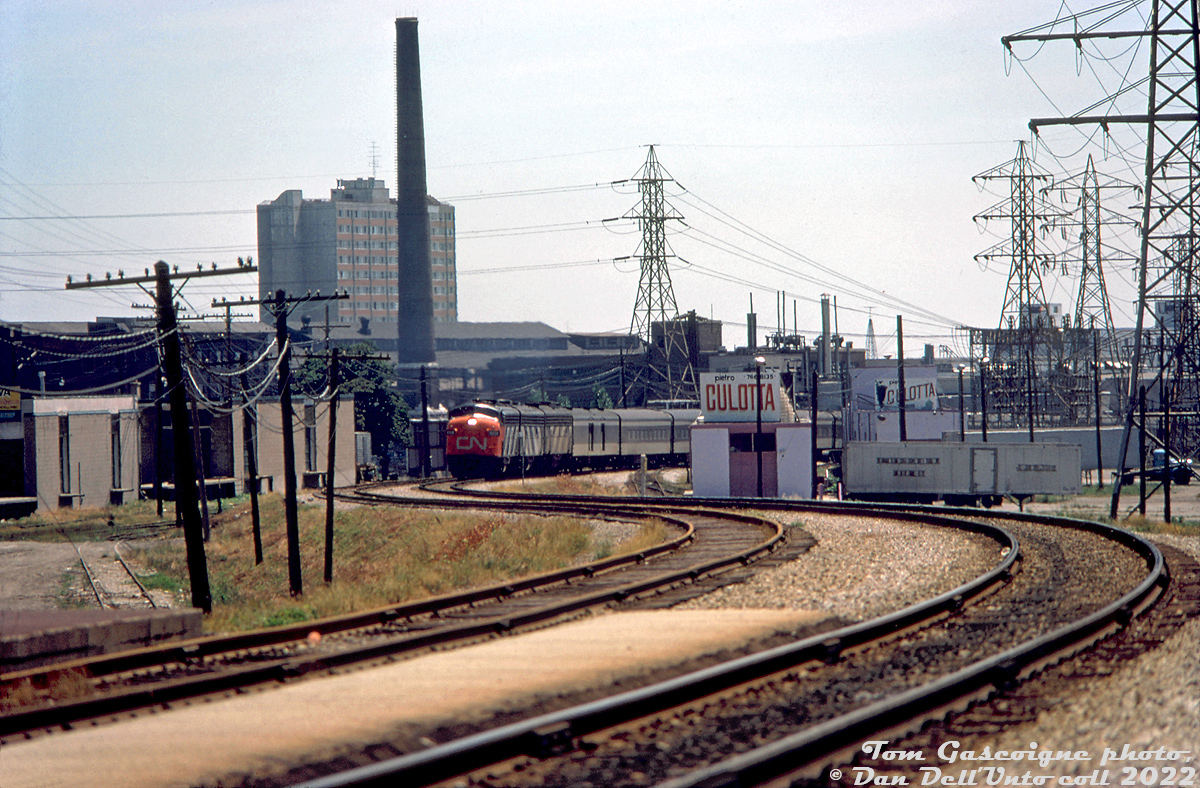 An A-B-B set of GMD F-units (lead by possibly CN 6526) heads northbound through mid-town Toronto with CN's Super Continental, snaking around the curves of the Newmarket Sub north of Davenport diamond on the approach to St. Clair Avenue station. Timetables suggest this is train #103 (formerly #3), that had departed Union Station downtown just after 3pm.

The section of track in the St. Clair/Davenport/Caledonia area here had been grade separated in the late 1920's/early 30's, that coincided with demolition of the old Davenport Station and construction of a new brick St. Clair Avenue Station. Visible in the background is the large headhouses of the Canadian Generel Electric (ex-Canada Foundry) Davenport Works plant. Power lines are from Toronto Hydro's nearby Witshire transformer station.

Due to its proximity to Little Italy, various food and produce distributors established buildings here to receive boxcars and reefers of fresh produce, grapes, etc, sometimes from as far away as California. On the right (west side of the tracks) are Pietro Culotta Grapes, which had two buildings with multiple sidings and loading platforms. Out of frame, A&P had a distribution warehouse nearby and sidings coming off the same spur. On the left (east side) of the corridor are Caledonia Food Distributors (pictured, note loading doors along siding), Darrigo Brothers, and the Meschino Banana Company. CN also had team tracks and a ramp track here.

Remnants of sidings and much of the old buildings here did survive into the late 2000's before progress and redevelopment took hold. The Newmarket Sub, single tracked through here at one point, will probably get double-tracked again once Metrolinx finishes its Davenport diamond grade separation project.

Tom Gascoigne photo, Dan Dell'Unto collection slide.