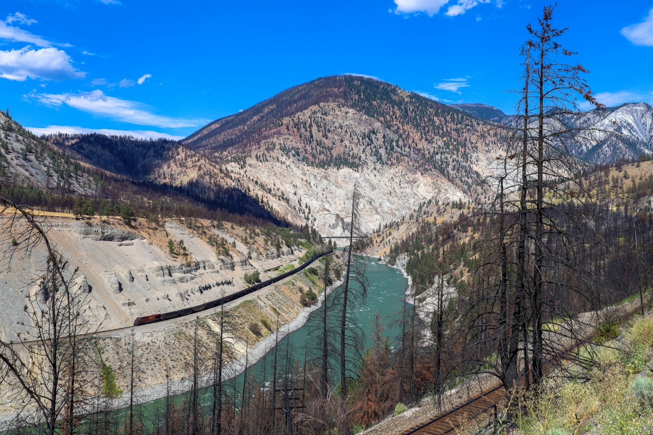 CN C 73151 01 eases towards the former townsite of Lytton, BC which was completely destroyed by a wild fire in 2021.  The spectacular White Canyon in the distance, along with the pristine blue Thompson River provides stark contrast to the fire singed trees and brush.