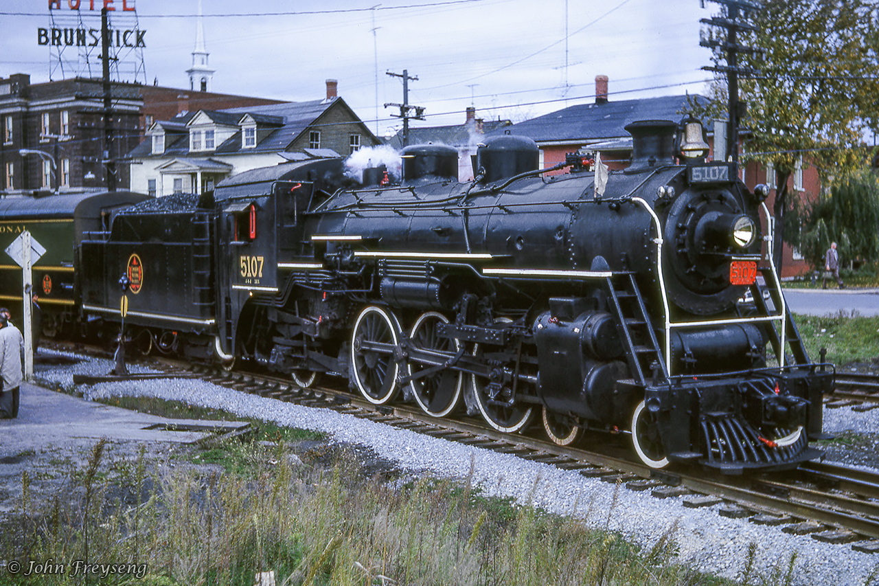 The weekend of October 13-14, 1962 provided railfans in the Montreal area with two excursions out of Central Station behind two steam locomotives. The Saturday trip would be led by J-4-d Pacific 5107, and the Sunday trip double headed with 5107 and U-2-c 6153. These trips run by the Canadian Railroad Historical Association were supported by the Upper Canada Railway Society.CNR 5107 is seen sitting to the north of CN's Richmond station during a water stop while en route from Montreal - Sherbrooke. The six car train had departed Central station track 9 that morning at 0829h making a water stop at St. Hyacinthe and three photo runpasts. One more runpast would be held between Richmond and Sherbrooke before turning the train for the return to Montreal, stopping for three more runpasts and water on the way. Back in Montreal, 5107 would be cut off at Bridge Street, leaving the consist in the care of MLW S4 8062 for the last couple of miles into Central Station. Today the Sherbrooke sub is operated by the St. Lawrence & Atlantic Railroad, and CNR 5107 is on display in Kapuskasing. Bill Thomson caught 5107 at Mimico in the summer of 1958.Directly across from the station on Rue Principale Nord (Main Street North) sits the Hotel Brunswick. Built circa 1910, the hotel served many travelers over many decades. Like the former station, now a restaurant, the building serves a new purpose today, as the Résidence Brunswick, a retirement home. Michael Berry's 2015 shot across the Richmond yard gives viewers the station at centre with the Brunswick off to the left.Scanning and editing by Jacob Patterson.