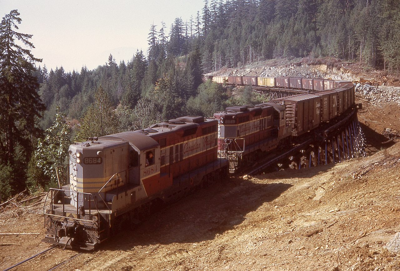 On Vancouver Island, CP’s line from Parksville to Port Alberni was ruled from 1949 to 1971 by Baldwin diesels, but increasing tonnages from the mills in Port Alberni plus the risk of fire from the common steel brake shoes on prolonged grades resulted in trial of pairs of GP9s with dynamic braking.  At the same time, due to advanced age of two large timber trestles, diversions on culverted fills were constructed.  This is an eastward train powered by 8684 and 8649 on the Four Mile Creek bridge at mileage 24.4, just east of the siding at Stokes.  In another half mile, bridge 23.9 over Five Mile Creek will be encountered.

Stokes, in common with numerous other stations on the Port Alberni sub., is named for one of its surveyors, this one for William Marpole Stokes, later an E&N division engineer.