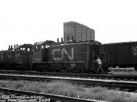 CN SW1200RS units 1204 and 1208 (1204 the first CN SW1200RS built, and both part of CN's first order delivered in 1956) switch Danforth Yard in Toronto's east end in June of 1980. A crewmember hops on the rear unit's foodboard as the pair moves away, past an old 40' boxcar bearing the old maple leaf "wafer logo". The apartment buildings in the background at Main St. & Danforth Avenue were probably some of the first that sprung up in the area, built on former coal yard and lumber yard property in the early 1970's (that may have caught fire before then).<br><br>Danforth Yard was CN's main east-end Toronto yard (smaller than the main west-end Mimico Yard). Equipped with a roundhouse and steam servicing facilities, it was at the top of a helper district from Union Station that often saw large 4100-series 2-10-2 steamers assisting heavy trains on the 1.2% grade climbing eastbound out of Toronto. A good number of lumber, coal and aggregate industries that used rail service were also located around and nearby the yard in this part of Scarborough. There was also, of course, a CN station here, and later a GO Transit station (the old CN station building was demolished in the mid-70's).<br><br>Danforth Yard's importance declined over the years as steam transitioned to diesel, industry moved out of the city, and freight traffic was shifted around Toronto proper to MacMillan Yard to the north. By the 80's Danforth was basically used for MofW car storage and repairs, and also had a bridge repair shop on the property. Those operations were closed in 1986, and the yard was eventually removed and sold off for redevelopment in the late 90's. Little remains today except for a GO station and some local railway-themed street names.<br><br><i>Bill Grandin photo, Dan Dell'Unto collection negative.</i>