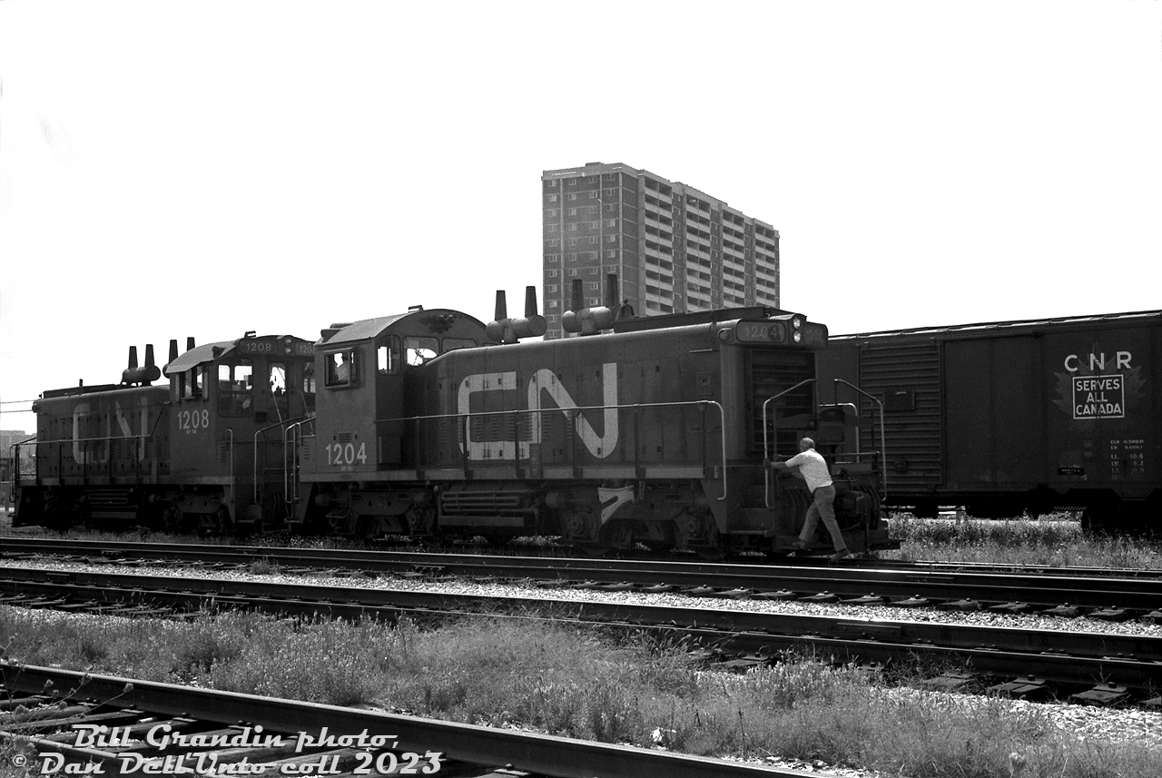 CN SW1200RS units 1204 and 1208 (1204 the first CN SW1200RS built, and both part of CN's first order delivered in 1956) switch Danforth Yard in Toronto's east end in June of 1980. A crewmember hops on the rear unit's foodboard as the pair moves away, past an old 40' boxcar bearing the old maple leaf "wafer logo". The apartment buildings in the background at Main St. & Danforth Avenue were probably some of the first that sprung up in the area, built on former lumberyard property in the early 1970's (that may have caught fire before then).

Danforth Yard was CN's main east-end Toronto yard (smaller than the main west-end Mimico Yard). Equipped with a roundhouse and steam servicing facilities, it was at the top of a helper district from Union Station that often saw large 4100-series 2-10-2 steamers assisting heavy trains on the 1.2% grade climbing eastbound out of Toronto. A good number of lumber, coal and aggregate industries that used rail service were also located around and nearby the yard in this part of Scarborough. There was also, of course, a CN station here, and later a GO Transit station took its place.

Danforth Yard's importance declined over the years as steam transitioned to diesel, industry moved out of the city, and freight traffic was shifted around Toronto proper to MacMillan Yard to the north. By the 80's Danforth was basically used for MofW car storage and repairs, and also had a bridge repair shop on the property. Those operations were closed in 1986, and the yard was eventually removed and sold off for redevelopment in the late 90's. Little remains today except for a GO station and some local railway-themed street names.

Bill Grandin photo, Dan Dell'Unto collection negative.
