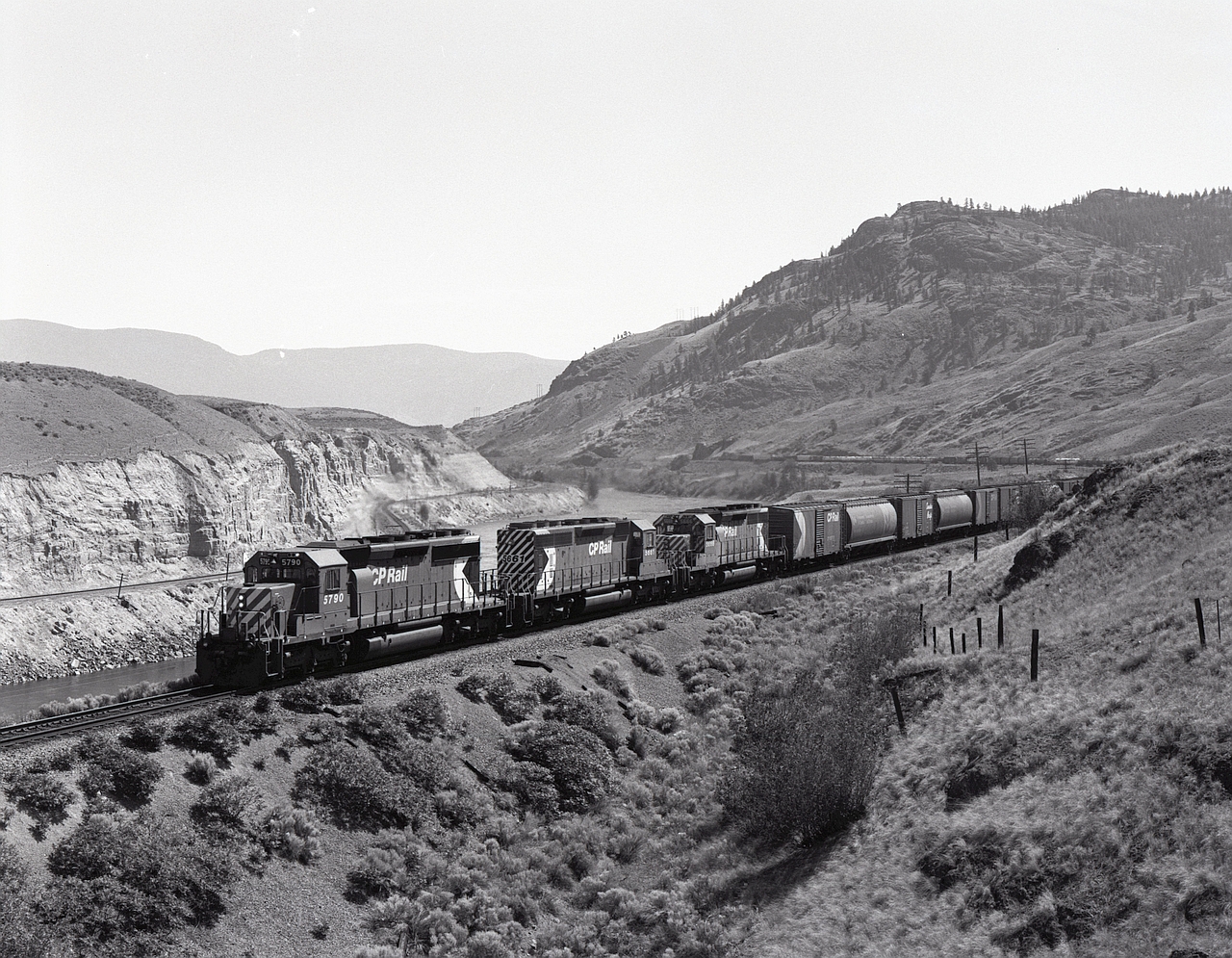West of Kamloops, CP follows the south shore of Kamloops Lake to Savona, then starts a significant upgrade along the Thompson River to Walhachin.  In earlier years, this was a helper grade with daunting steeper sections instead of today’s steady climb.  On Sunday 1980-06-08 at 1107 PDT, a westbound with SD40-2s 5790 + 5661 + 5666 (one with wide stripes and two with narrow ones, and all with full-height multimarks) is working hard at mileage 28.5 with three more miles to Walhachin.

In the distance near the tail end, evidence of the lower older grade can be seen, and on the other side of the river is the CN Ashcroft sub.