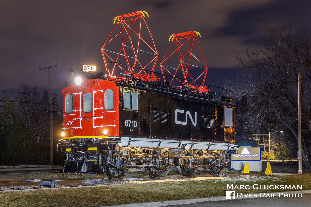 Repainted and Relighted. When former Canadian National Railway Company 6710 (Z-1a, General Electric, 1914) was retired from service in 1995, it was donated to the Village of Deux Montagnes, Quebec, then the end of the Deux Montagnes Line. After sitting as a stationary display for a number of years, it became eroded by the elements. In 2019, it was moved to Parc Belair about a mile from that location. The locomotive was repainted and even the pantographs now reach upwards towards an invisible catenary again and the headlights and number board shine brightly at night, as seen on Monday, April 3, 2023. The line is currently closed while it is being reconfigured for light rail operation.