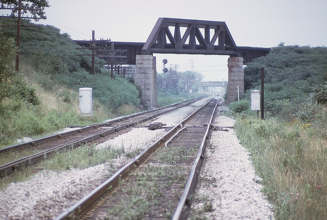 It has been at least 8 years since on this site there was a discussion about the CN Thorold Sub north track bridge over the CN Grimsby sub down by the canal at Merritton. Knew I had a photo somewhere. Finally found it.
The Thorold Sub used to be a two track main, and pictured is the north track bridge as it went over above the Grimsby and down into the yard at Merritton.  The bridge was removed in 1976 although some of the track on the north side leading up is still there. The Thorold sub used to be  busy line until the Canal Bypass was built in 1968-70 and what used to be the Welland sub became the Stamford and a connection was put in at CN Clifton, bypassing the steep grades at Thorold.
I had been told that this bridge was known locally as "Bum Bridge" as it was a hangout for hoboes and others hopping trains and was the source of many injuries due to reckless behaviour of those who frequented the area.  So the bridge is long gone but part of the abutments can still be seen.  In this image looking west the background visible is the Merritt St overpass and behind that, the old Merritton NS&T bridge.
Note in the foreground the canal 'derails'.