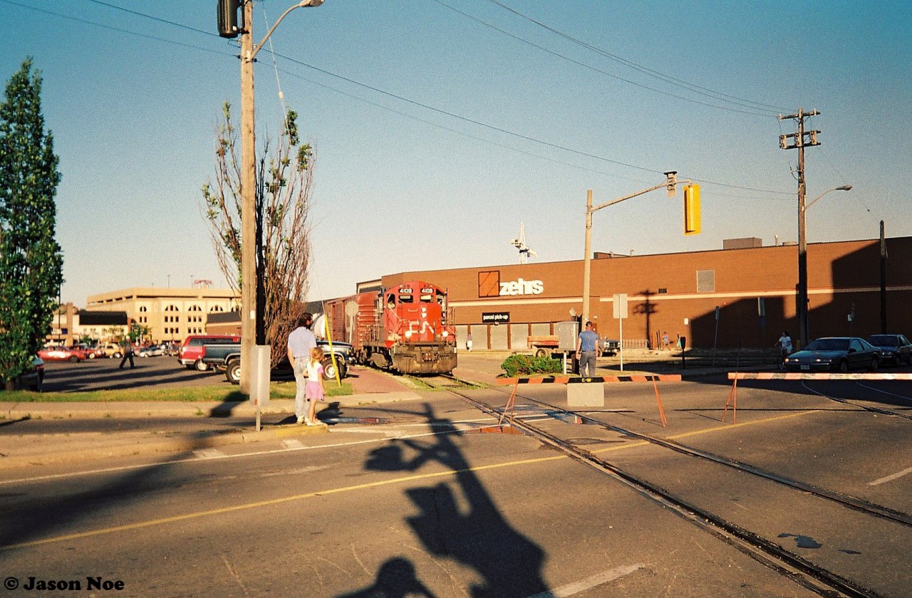 So the story goes, on July 12, 1993 a massive fire ripped through the Seagram’s distillery in downtown Waterloo, Ontario. The fire destroyed the original still-house and mill building that faced Erb Street West near the intersection with Caroline Street. Thankfully the Seagram Museum building was not damaged in the blaze. 

Later that evening, CN’s 15:30 Kitchener Job with GP9RM 4109 is seen slowly making its way through Waterloo Town Square on the Waterloo Spur as they headed to Elmira. Conductor Rollie is keeping a watchful eye as they approached the fire scene and two barricades at the intersection of Erb and Caroline Streets. To the right of the Waterloo Spur is the CP Waterloo Subdivision, which saw CP making their last run to Waterloo the week prior on the evening of July 6. The times were certainly changing in the city during that summer.