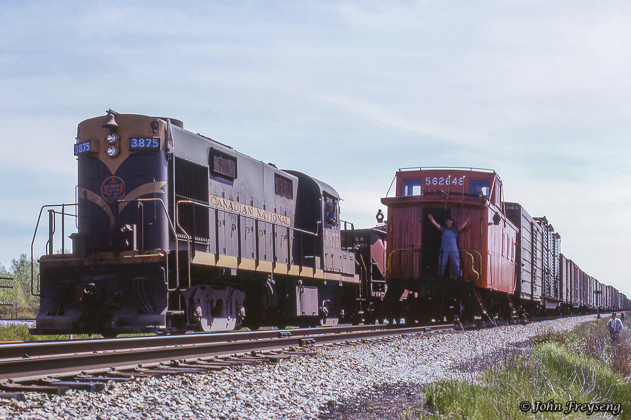 CN extra 3875 west sits in the hole at the west end of Canfield siding waiting on an eastbound Norfolk & Western freight to clear.  The friendly tail end crew member on the rear of caboose 562648 gives a wave to both crew and railfans trackside.Scan and editing by Jacob Patterson.
