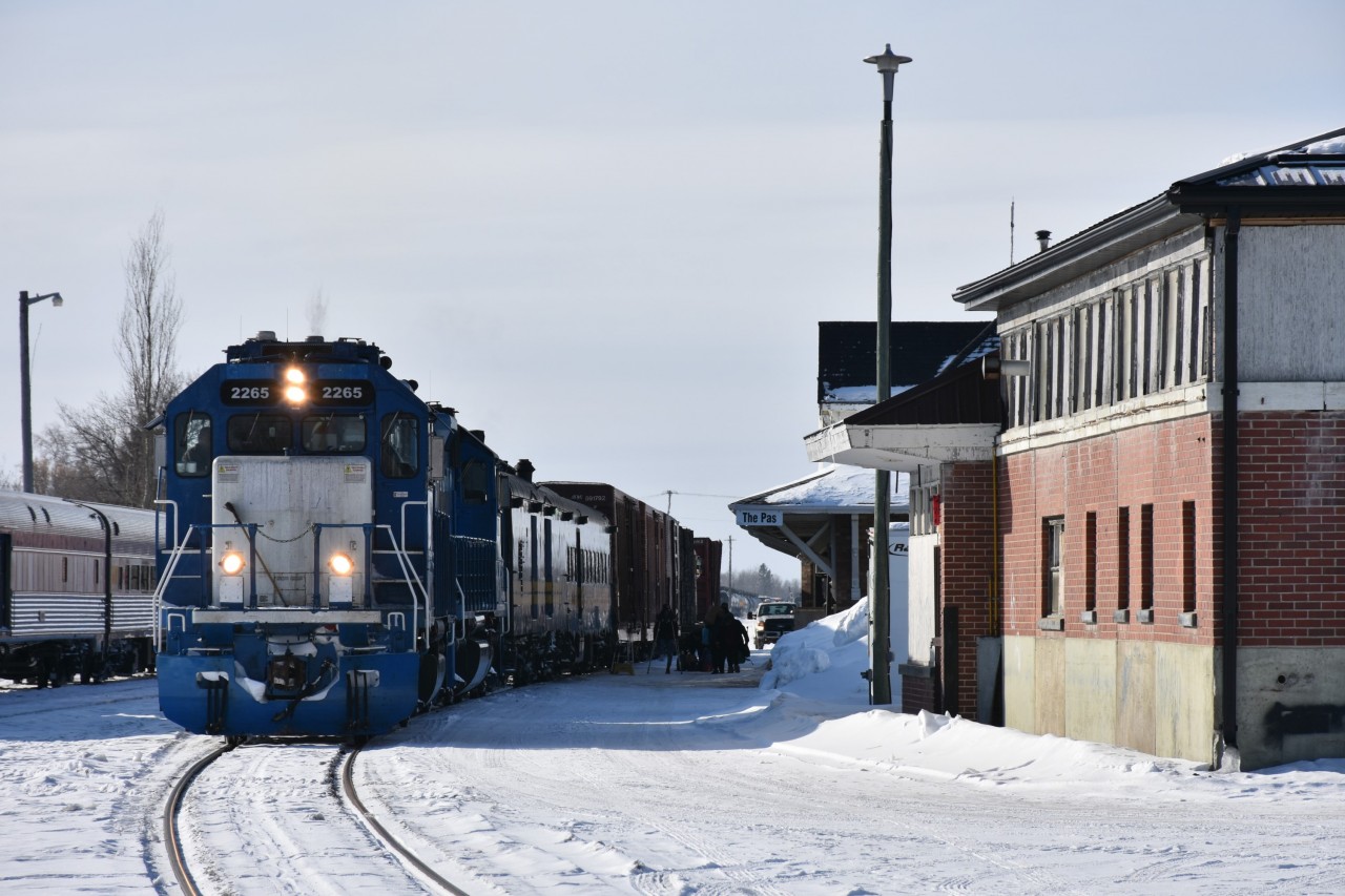 Three different railways are represented in this photo at The Pas, MB. on this blistering cold March 16, 2023 morning. 
At the left of the frame is VIA Rail's #691 consist of stainless steel Budd built passenger equipment waiting to return to Churchill, on the main in front of the station is KRC's (Keewatin Railway Co.) #291 The Pukatawagan Mixed with classic ex-VIA, exx-CN baggage car and coach loading up with passengers, baggage, and supplies, and, the red brick building to the right is HBR's (Hudson Bay Railway) Maintenance building at 313 Hazelwood Ave. The Pas. 
The only remnants identifying this location as a former Canadian National northern Manitoba hub are three old zebra striped locomotives (renumbered for HBR) sitting stored out of sight in the distant yard near the roundhouse/diesel shop facility.