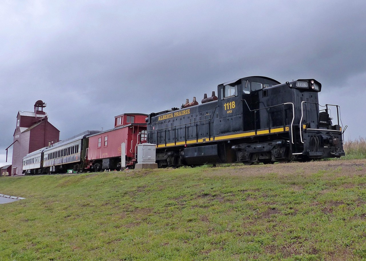 The locomotive is APXX 1118(GMD1) formerly CN 1118(GMD1) built in 1958 and currently in use by Alberta Prairie Rail Excursions. The caboose is APXX 79146. In the background is the former P&H grain elevator that was built in 1920 and closed in 2003. Currently it is a museum run by the Stettler P&H Elevator Preservation Society.