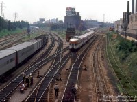 Hazy day in "The Big Smoke": the Maple Leaf billboard reads 26 degrees as switch tenders in charge of the manual ground-throw interlocking switches watch the tail end of CP train #11 "The Canadian" (Toronto-Sudbury section) departing downtown Toronto at Cabin D, leaving Toronto Terminals Railway trackage onto the start of the CP Galt Sub near Tecumseh St. tower. Today's train is a long summer consist, with power appearing to be two F-units bracketing an RS10. A silver-painted 2200-series steel coach or two can be seen near the head end. In the distance on the right is CP's Parkdale Yard, with the headlight of a switcher barely visible. The red farm equipment on flatcars on the right (off CN's Weston Sub) was from the nearby Massey Ferguson plants in the Liberty Village/Parkdale area.<br><br>On the left, a CN Tempo train powered by two RS18's (with one of the three HEP baggage cars) is seen departing west on the Oakville Sub. In the distance to the left, an inbound GO train approaches, with the tail end of an outbound train visible under Strachan Ave. bridge (possibly stopped at Exhibition GO Station). Also next to them is a CN switcher working the Bathurst South Storage yard.<br><br>The usual Bathurst Street 70's set pieces are in attendance:<br>-Cabin D interlocking tower presiding over the manual interlocking (tower removed and location renamed "Bathurst Street" when the interlocking plant was rebuilt/automated around 1983, remotely controlled from John Street tower)<br>-CP's Tecumseh St. tower further back (controlled movements from CP's Galt Sub & Parkdale Yard across CN's Weston Sub to the Simcoe St. freight shed lead on the far right).<br>-Steam-era "tell-tales" strung west of the bridge to warn any crew members riding the tops of freight cars of the Bathurst St. bridge (Bathurst North Yard behind us was still a CN freight yard at the time).<br>-The ever-prominent Inglis appliances sign facing the Gardiner Expressway, and the sprawling buildings of the old Inglis plants and other industrial buildings of Liberty Village (today, all mostly gone or converted into condos/lofts, dwarfed by more and more new condos).<br><br>Bathurst Street is still as busy as ever today, but about the only thing that hasn't been changed, added, removed or upgraded today is the fact that it's still a rail junction!<br><br><i>Keith Hansen photo, Dan Dell'Unto collection slide.</i>
