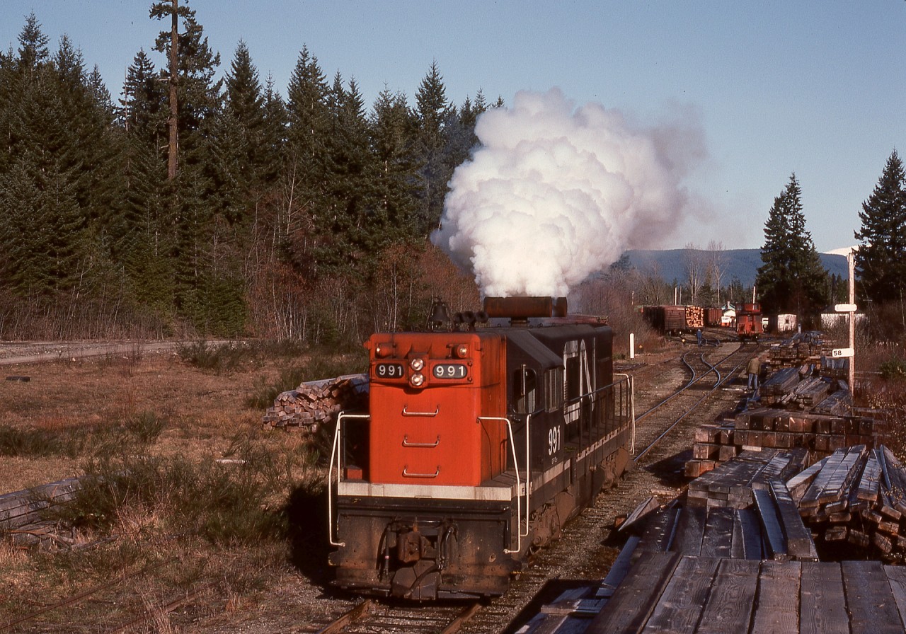On Vancouver Island, Thursday 1976-04-01 was frosty, and G12 CN 991 was left idling overnight at Deerholme to prevent freeze-up, leading to an excessive accumulation of exhaust system deposits.  Well aware of the unpleasant smoke likely to affect the tailend crew, the engineer worked the engine against full-set engine brakes to “clear its throat” on a short run eastward before tying onto the distant pole loads and caboose for a run down to the barge slip at Cowichan Bay.