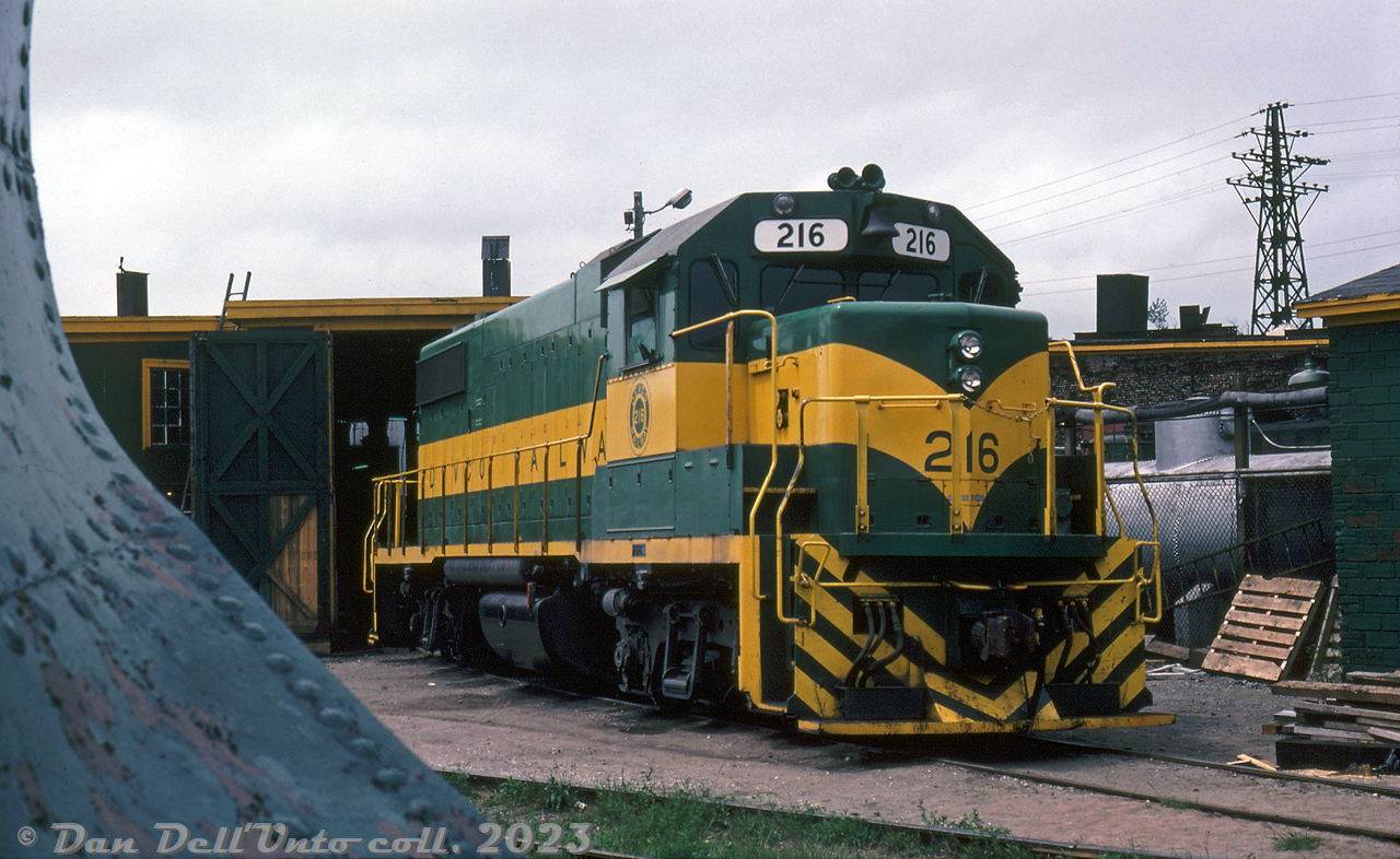 DEVCO Railway 216, a GMD GP38-2, is pictured at their Glace Bay shops partially framed by an old steel wedge plow parked nearby. This unit was part of an order of four units, 216-219, built at GMD London in May-June 1979, and delivered together in early July.

This order was unique in that owner DEVCO (Cape Breton Development Corp) specified these four units be equipped with 575 volt, 1000 kilowatt generators inside them to cover for emergency power outages. The electrical plugs for outputting 3-phase electrical power are visible on the right side of the short hood. The unit survives today as NBSR 2317.

Original photographer unknown, Dan Dell'Unto collection slide.