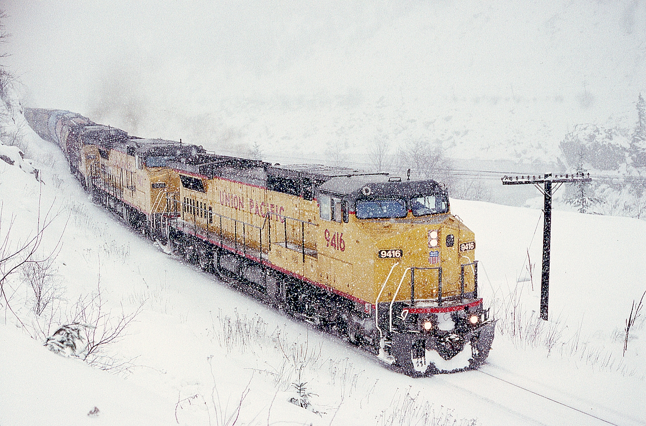 Directly below the Saddle Rock Tunnel on the Trans Canada Highway in the Fraser Canyon, a CP grain train with three UP Dash8-41CW units on loan for evaluating possible General Electric locomotive purchases (which eventually occurred in 1995) is experiencing winter snow as they leave Saddle Rock at 1503 PST on 1991-03-03 headed for Yale and onward to the CP yard in Port Coquitlam.

A slightly later view posted a while back is at .