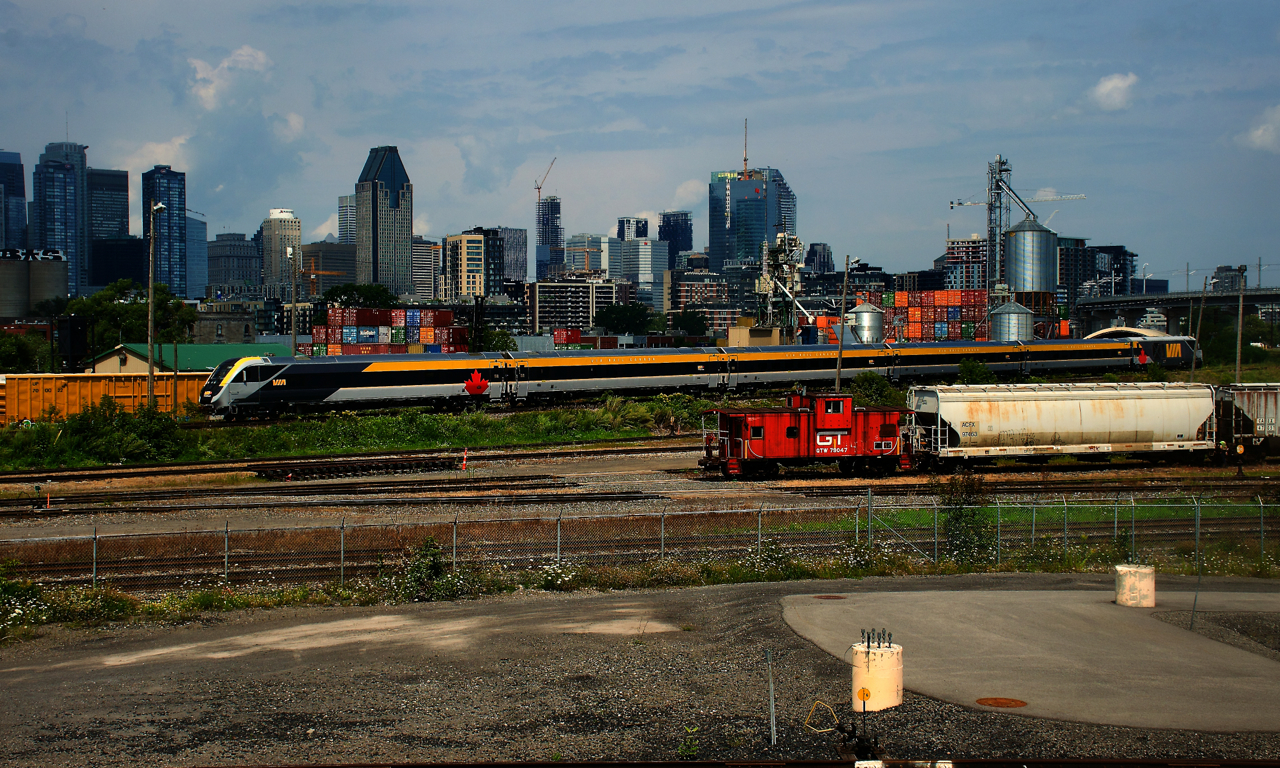 VIA 35 has a Siemens trainset as it passes the skyline of downtown Montreal. A GTW caboose is seen at the end of some cars in the Pointe St-Charles Yard.