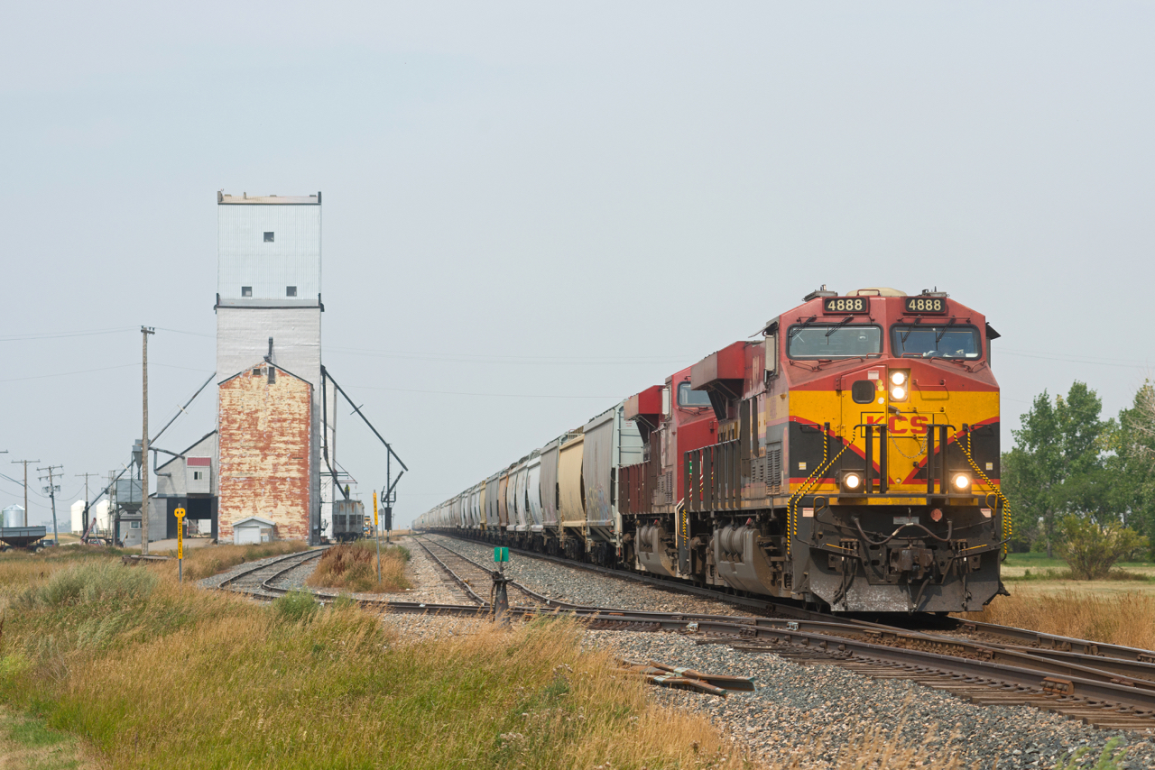 With a hazy sky due to wildfire smoke, a loaded CPKC grain train makes its way south along the Weyburn Sub towards the US border. Up front is Kansas City Southern de Mexico 4888, an odd sight along side the old pool grain elevators of Lang Saskatchewan.