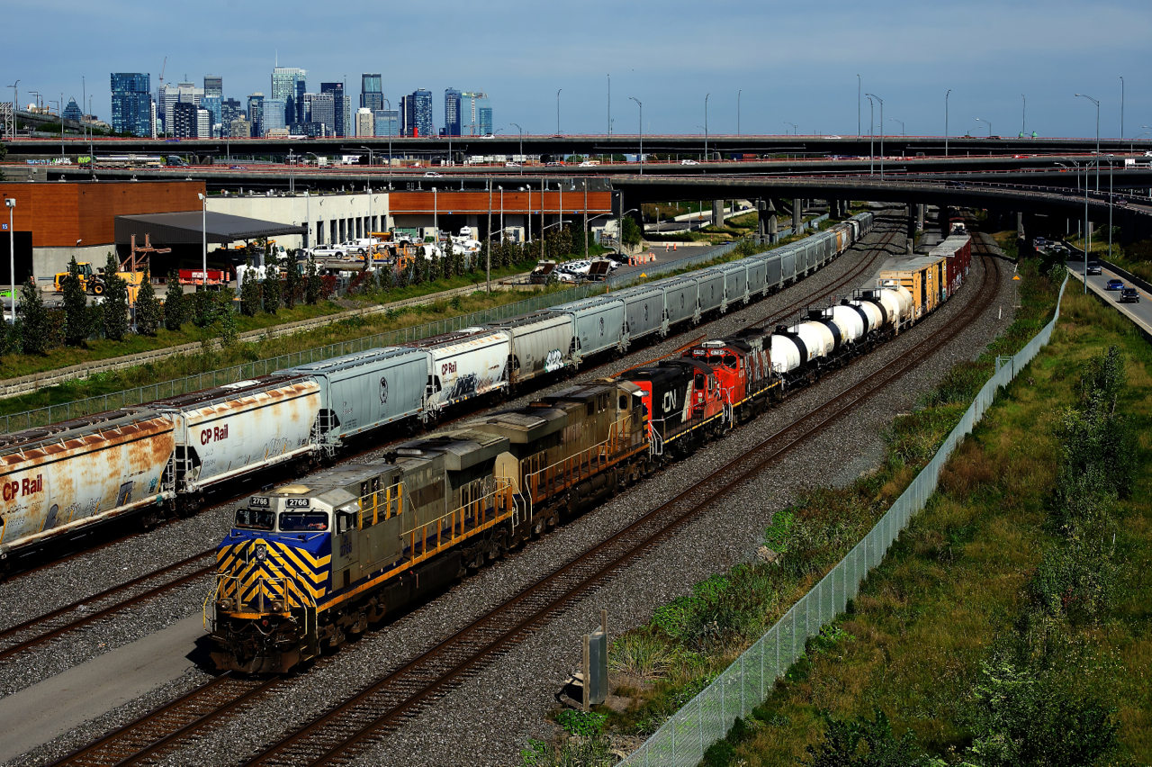 A 588-axle long CN 527 has two ex-CREX units followed by two geeps as it passes the skyline of downtown Montreal. At left is a long line of CP grain cars, for various clients in Montreal located on CN lines.