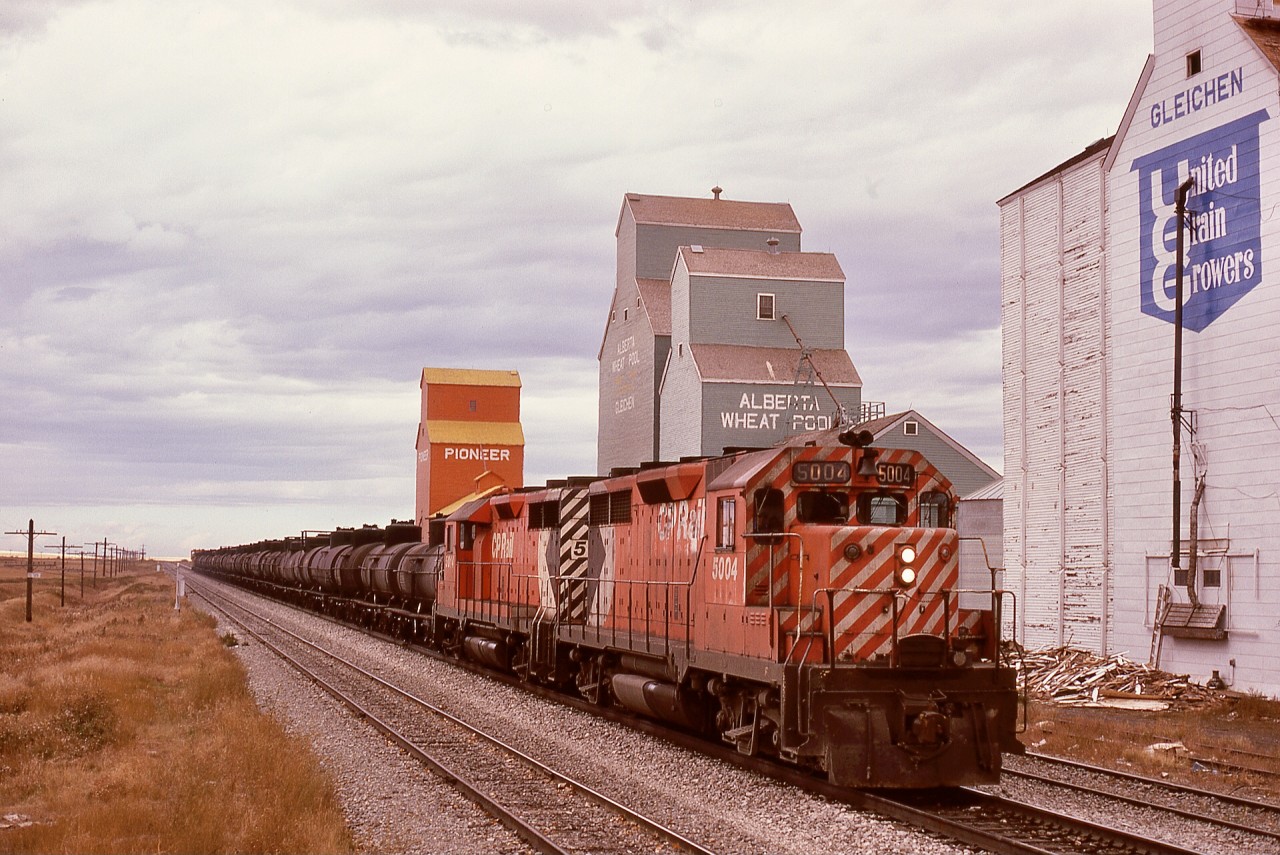 From Alyth yard in Calgary, CP ran a Princess Turn eastward 75.7 miles on the Brooks sub. mainline to Bassano then eastward 41.7 miles on the Bassano sub. to an oil loadout at Princess.  On Wednesday 1974-10-02, that job was running as timetable train No. 96 with a pair of GP35s, 5004 and 5014, as it passed milepost 125 near the grain elevators at Gleichen, with 27.2 miles to reach the junction at Bassano.

In my ignorance, assuming a turbocharged GM was an SD40, those similar-sounding GP35s were entered into my notebook as 5504 and 5514, with that error not corrected until I got my slides back from that trip!  If only I had known those units would be my exasperating work when they were assigned to CP’s (Port) Coquitlam locomotive facility ten years hence.