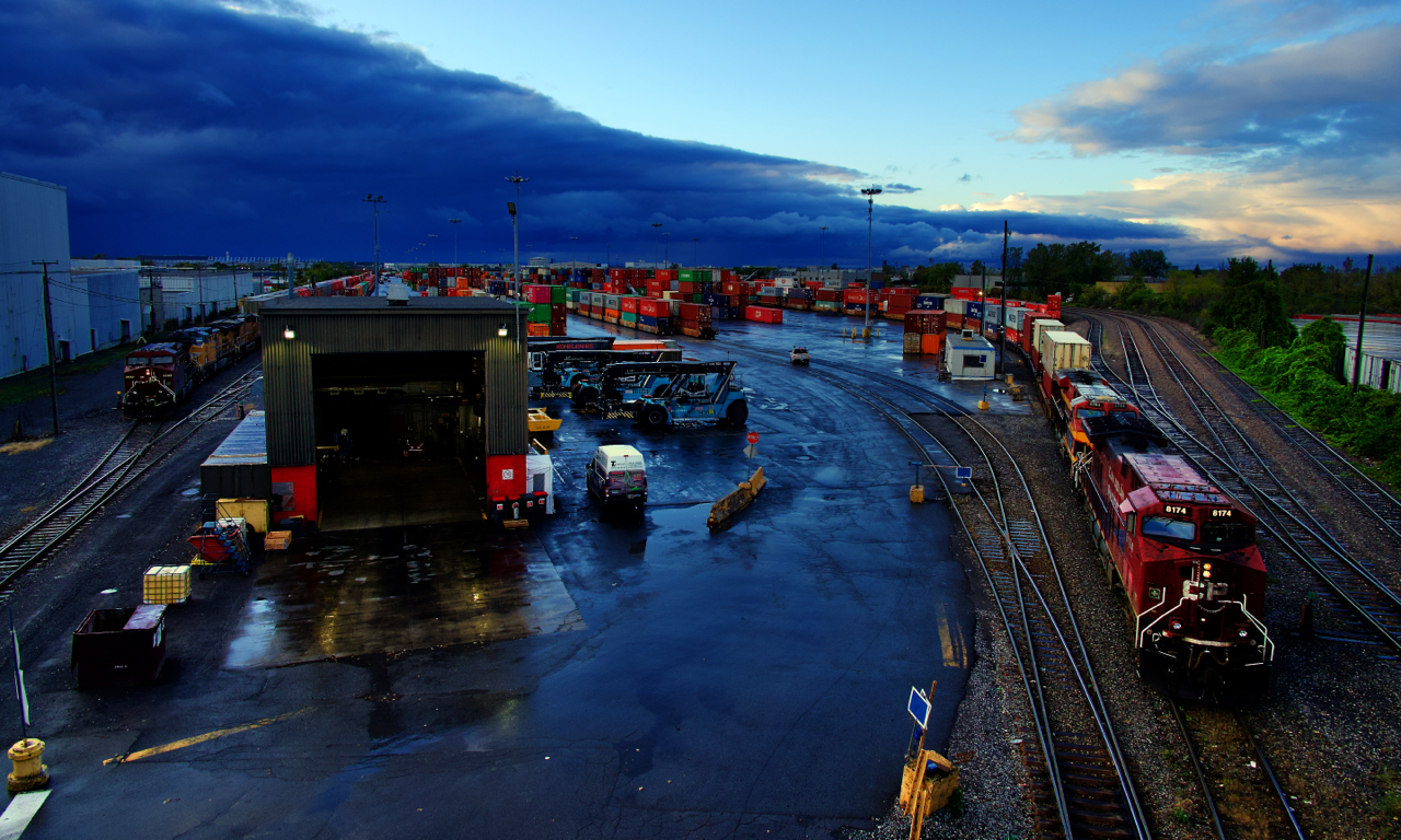 A dramatic sky is overhead as uncommonly two trains are in Lachine IMS Yard simultaneously. At left CPKC 112 is waiting for CPKC 119 to finish its lift before heading to St-Luc Yard with its power and autoracks.