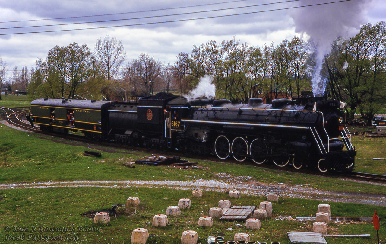 After arriving at Cobourg and cutting off from its train, CNR 6167 shoves the baggage car of fans around the wye in preparation to return west to Oshawa, and Toronto Union Station.  Today's excursion included a stop at Oshawa for traction fans to take in the Oshawa Railway operations which were in the last few years of service. This day marked the last train down King Street, celebrated by a parade as motor 300 hauled a single boxcar through town.Once around the curve, 6167 will be on the remnants of the Cobourg & Peterborough Railway, built in 1853.  The line later came under control of a few small companies and ultimately the Grand Trunk Railway before abandonment circa 1898.  A small portion is still in use to serve the Canada Pallet Corporation about a quarter mile away from this scene.  Additionally, in the grass area beyond the baggage car, just above the conductor, the former CNR Orono Subdivision, originally the Canadian Northern Railway Toronto - Ottawa mainline, crossed this scene until abandonment in 1923/1924.  The spur leading in that direction used a very short stretch of the old main to access a few industries east here that have since stopped using rail.Steve Bradley would shoot the special after its westbound departure from Oshawa highballing through Port Union.Tom J. Gascoigne Photo, Jacob Patterson Collection Slide.