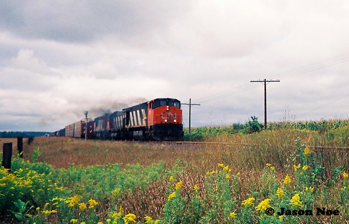 Back in September 1993 CN completed a large-scale bridge replacement west of Paris on the Dundas Subdivision. This resulted in CN rerouting trains not only on their Guelph Subdivision, but also detouring across Canadian Pacific’s Galt Subdivision for several days. In the west, the trains would begin their journey to the Galt Subdivision through the interchange track at Woodstock then onto the CP St. Thomas Subdivision for a short stretch before reaching the Galt mainline.

During one of those detour afternoons, I left school early and my dad and I drove to Wolverton to hopefully catch a CN train on the CP Galt Subdivision. We were heading to Woodstock but made a brief stop as this had become one of our favorite spots to railfan at the time. If memory serves we actually didn’t have to wait long until the scanner crackled and a headlight came into view from the west. 

Here CN 410 highballs through the east siding switch Wolverton with 2111, 2320 and 2117 just as the sun pierces a small hole through the late summer overcast. I remember clicking the shutter of my small Kodak camera as fast as I could to hopefully capture something. More than 30 years later I would say I was able to get one that was semi-decent.