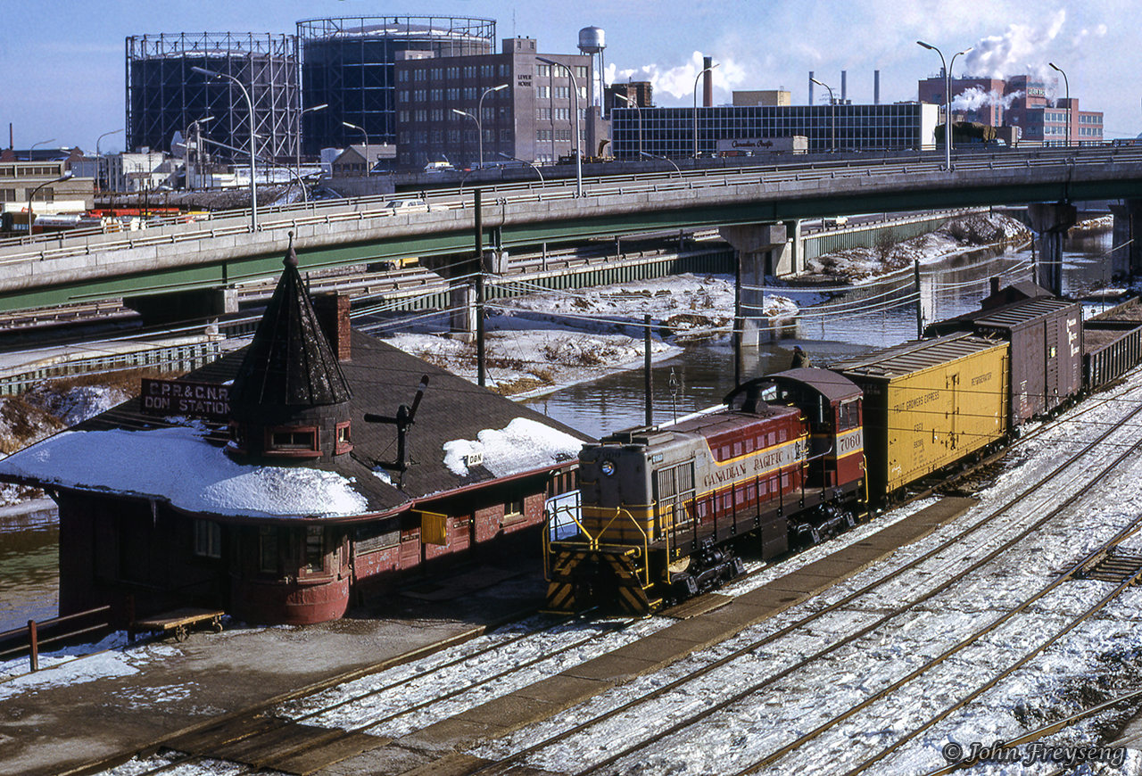 CPR 7060 leads an eastbound transfer from Parkdale Yard up to Leaside, taking orders at Don Station. With its two-position train order signal, showing only proceed and stop, the yellow flag (lantern by night) has been hung beneath the order board allowing the orders to be handed off on the fly. A great book, "OS Don" by John Mellow, details operations around this station very well. Get your copy through the Bytown Railway Society.Looking across the Don River, we can see the Lever Brothers' Soap Factory, built in the 1940s and currently under demolition after failed heritage adaptive reuse petitioning, and farther beyond, the gasometers at Consumers Gas Station B. Station B closed by 1956, with the gasometers being demolished in 1969. Note the Canadian Pacific Express trailer above on the Eastern Avenue Diversion bridge.Scan and editing by Jacob Patterson.