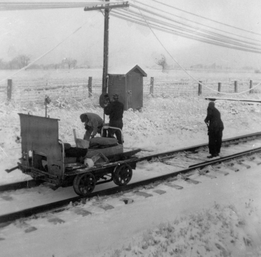 All in a winters day work  
The section guys are out completing their daily winter chores. A freshly cleaned and filled lamp being set atop the switch stand, as the switch points and throw rod get a good clearing of accumulated snow. 
The hanging wires suggest a heavy wet snow has covered the area which will keep this 3-man crew and foreman quite busy on their territory this cold winter day.