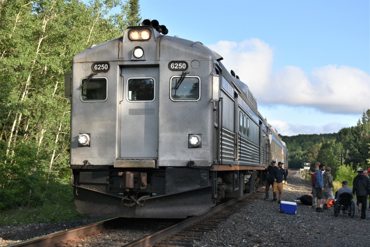 Plain Jane Budd  
VIA #186 the Sudbury to White River train has stopped at Mile 88.8 on the CPKC White River Sub. to detrain and entrain passengers going to and coming from the famous Lodge 88 camp which is accessed from this location. 
There's lots of activity on the ballast shoulder and right-of-way as camp supplies and customers gear is loaded and unloaded from the baggage section of the leading Budd car. 
The nose of VIA 6250 is as plain as it gets! Basic silver/grey paint, no VIA logo, no reflective striping, no stenciled running number.