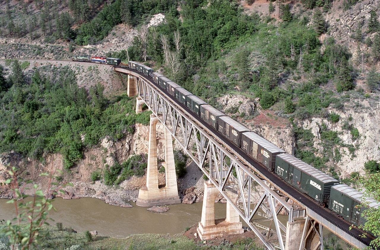 With a mix of box and empty wood chip cars, four BC Rail  SD40-2's; two mid train, crossing the Fraser River, 


 North of  Lillooet, B.C., near  Mile 155  Squamish Sub.,


May 25, 1989 Kodachrome by J. Art Clowes, collection of S. Danko


The Location:


Searching through google maps aerial photos, and based on the bridge design,  Lillooet appears to the closest match – would appreciate someone in the know to confirm.
 

Noteworthy


In 2004 CN bought  BC Rail Ltd., the assets of the operating railway (the loco's, rolling stock).


And in 2004 CN leased the British Columbia Railway Co. (BCRC) assets (excluding the Delta Port assets) including the right-of-way and track materials, for a period of 60 years. Per the terms of the lease, the rail, ties and ballast are owned by BCRC as would be any replacement materials, which must be of the same standard as the original materials, unless the landlord consents otherwise. Also at the end of the lease the assets must be at the same standard as at the start of the lease – which may be interesting especially with regard to the Dease Lake extension the 202 mile Takla Sub-Division (Fort St. James to Minaret end of track).


The outcome of the above was a significant political scandal, whereas other parties bidding for the assets claimed the process was rigged. Quite the kerfuffle ensued. In 2010 two BC government political aides were convicted of breach of trust  and sentenced  serving two years in house arrest and 150 hours of community work.


sdfourty