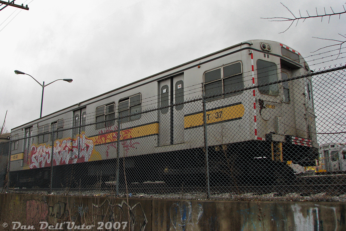 TTC G1 subway car RT-37, one of the last of Toronto's "Red Rocket" Gloucester subway cars still on the property in 2007 (built as TTC 5069 for Toronto's first subway line in 1953-54, retired by the early 90's), sits stored at the back of Greenwood Yard awaiting disposal. At the time, four of the "G's" remained, having been converted after retirement for maintenance of way use as power units for the railgrinder trains that profiled the rails around the subway system during off-peak hours. The sets were made of two G's for propulsion, bookending two small yellow railgrinder units towed in between. A yard collision in 2004 resulted in one set being retired, while the other set remained active until 2008. Due to their obscure duties during off-peak hours, they weren't very well known or photographed.

The story behind the hunt for these cars is somewhat long-winded, so viewers may want to go back to admiring the photo of RT-37 still in existence in 2007. It all started in December 2006, when someone on a discussion board posted a CP24 news article of some Oshawa GO line rider complaining about unsightly graffiti on subway cars parked next to the tracks hurting their eyes every day. Someone pointed out the blurry photo included, taken out of a rainy GO bilevel window, appeared to be some of the old TTC Gloucester work cars, stored on a back track in the subway yard along the railway corridor.

More intrepid, adventurous photographers were interested, and a plan was hatched by two to go investigate and get photos of the last of the G-series subway cars, conveniently inconveniently parked there. After a meetup at Union Station, hijinks and shenanigans followed with a visit to Bathurst Street bridge for GO, VIA & CN, some streetcar photos at the hip West Queen West & Bathurst, and an escorted visit inside TTC's Russell Yard for some CLRV and vintage PCC streetcar photos. After that, a short walk up Greenwood Ave brought us to the doorstep of TTC's Greenwood subway yard.

Unlike streetcar yards, one can't just knock on the door of a subway yard and get the red carpet treatment for an escorted tour inside, so scouting outside was the plan. Walking north up the east side of TTC's Greenwood Yard, on the off-chance the G's were visible from the main street, offered no luck. At the north end was Oakvale Avenue, which afforded a good look south into the yard, with lots of T1 and H6 subway cars (but still no G's among them). Then, a detour through the nearby neighbourhood side streets on the west side only yielded high sound barrier walls, with not so much as a peep possible into the yard. Coming out on Jones Avenue, there weren't many options left other than shooting them from the nearby CN rail corridor (if they were still there). Access at Greenwood and Jones Avenues was limited and too conspicuous, but a savvy navigator back then knew lots of the side streets had many "convenient trackside access options" available for the locals walking their dogs and ne'er-do-well crowd engaging in unscrupulous behavior and the like (this was before the Metrolinx days of tall fences everywhere, which was no doubt big business for some lucky fence contractors).

Sure enough, a dead-end street nearby offered a short dog walk option to the back of Greenwood Yard, with two TTC G-cars parked along the fence immediately visible for all to see: TTC G1 RT-37 (ex-5069) and G2 RT-34 (ex-5102). At the time, there were two steel (former red, now painted maintenance grey) G1's left on the property, and two aluminum (unpainted, but weathered grey) G2's, all sporting yellow maintenance striping.  RT-34 had been involved in a sideswipe incident with a T1 in 2004, and was retired & parked with a visibly damaged cab. RT-37 was presumably the sister car, and was parked with it, both on a disused dual-gauge track at the back of the yard that was formerly the CN siding leading into the property.

Despite the fence, photos were had, including the heavily rebuilt RT-11 "Duncan's Dragon" tunnel clearance car stored on the same track. Nearby was the other railgrinder set with G1 RT-36 (ex-5068) and G2 RT-35 (ex-5103) parked by the heating building at the south-west end, but people working nearby and poor photo angles discouraged getting too close. Satiated with photos, a short walk back to Jones Avenue and a ride on the Jones bus lead us back to the Bloor-Danforth subway, and after a bit of H4 subway riding it was decided to call it a day, with the mission complete and all main objectives achieved.