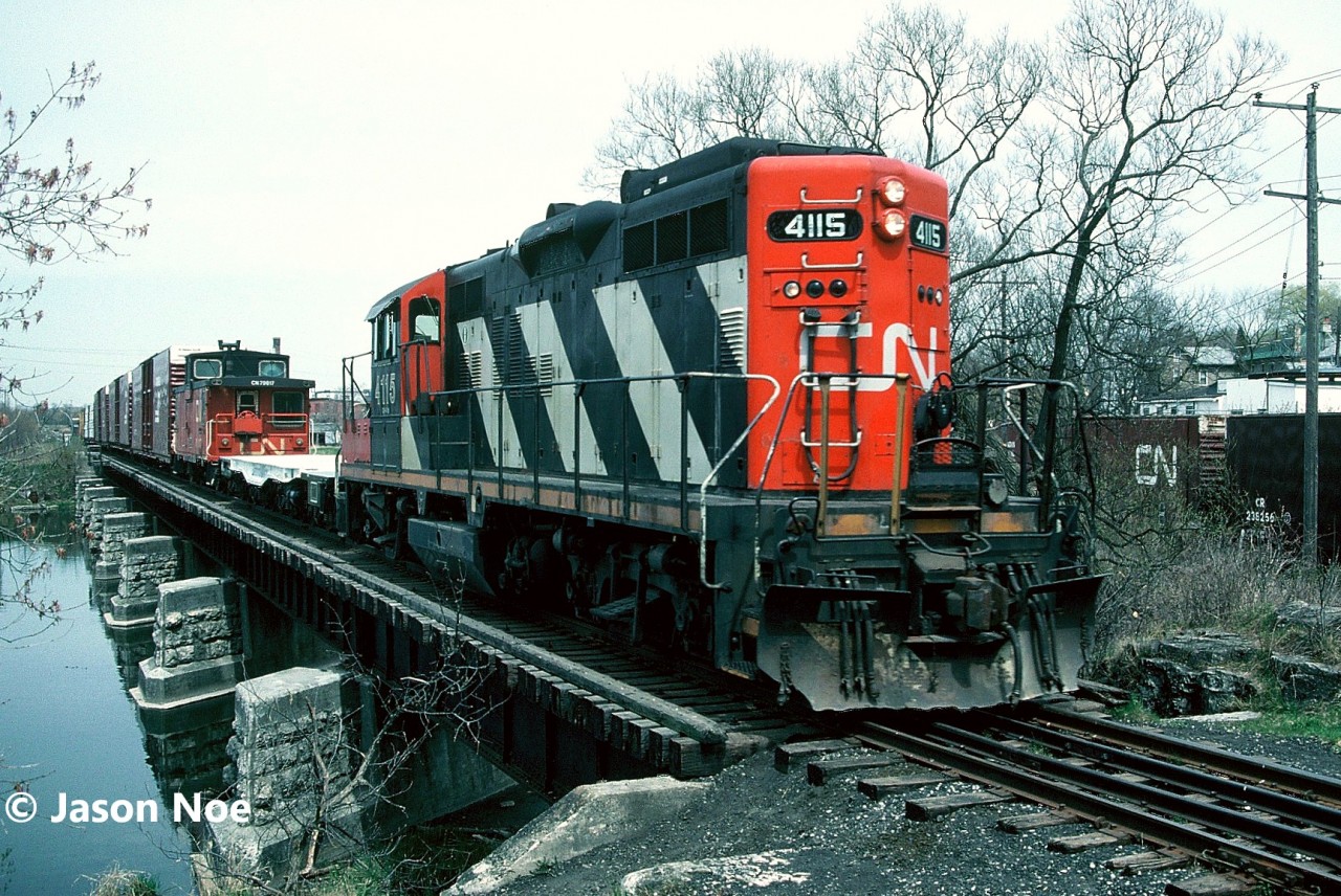 CN train 580 is pictured heading for industries situated on the Galt Industrial Spur in Cambridge, Ontario as it trundles over the Speed River bridge in the Village of Hespeler. Behind GP9RM 4115 is an empty dimensional flat that the crew will take to Babcock & Wilcox on the Fergus Subdivision later that afternoon. The auto parts boxcars to the right of the unit are sitting on the 1.2 mile Solaware Spur. 

This is a remnant of former Grand River Railway trackage that once operated through Hespeler, which was not far from where a station was located on the Hespeler Subdivision, subsequently becoming part of CP and downgraded to a spur. In later years, CN took over this trackage and renamed this section the Solaware Spur, and used it mainly to store auto parts boxcars for the various parts plants on the Galt Industrial Spur, including McPherson near Franklin Blvd.